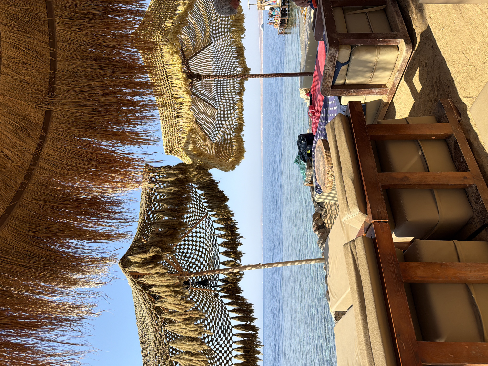 The image depicts a serene beachfront scene in Sharm El Sheikh, Egypt, with a clear blue sky and calm ocean waters. In the foreground, two thatched umbrellas provide shade over comfortable seating arrangements. The umbrellas are decorated with dangling tassels, adding a bohemian touch to the setting. Beneath them, cushioned loungers and wooden benches are arranged on the sandy ground, inviting relaxation. The sea stretches out to the horizon, with a distant shoreline visible and a few boats floating in the water. The overall atmosphere is one of tranquility and leisure, characteristic of a popular tourist destination.