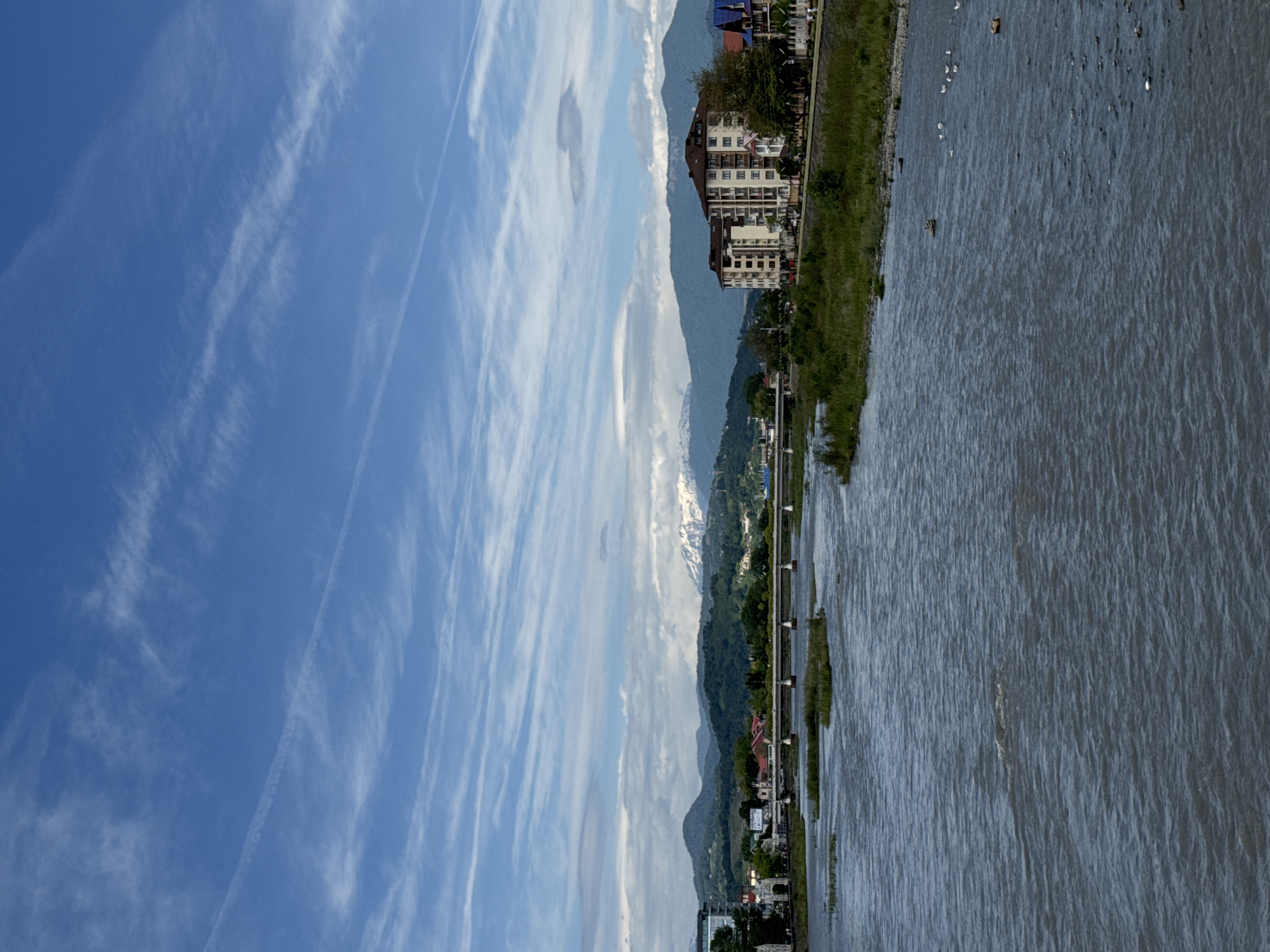The image depicts a view along the Mzymta River in Sochi, Russia. In the foreground, the river's surface is visible, reflecting the sky and surrounding landscape. The riverbank is lined with vegetation and a row of buildings, including a multi-story hotel. Behind the river, a mountain range is visible, with some peaks capped with snow. The sky above is blue with streaks of cirrus clouds.