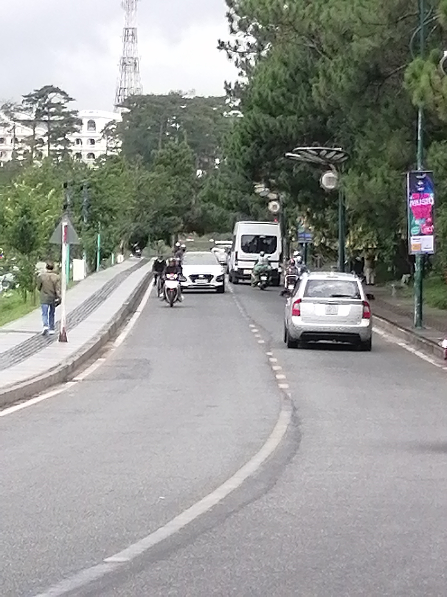 The image shows a street in Da Lat, Vietnam, with a white building and telecommunications tower visible in the background. A man walks along the sidewalk on the left, while a motorcycle, white sedan, white van, and a silver station wagon are traveling on the street. Trees line both sides of the road, and a street light with a banner is on the right side.