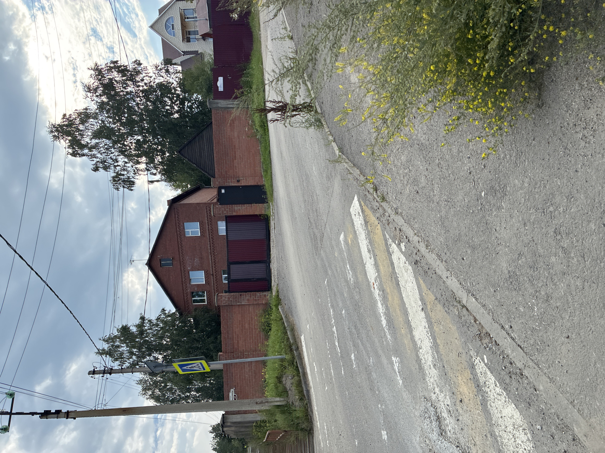 The image shows a street scene in the city of Markova, Russia, with a focus on a pedestrian crossing and the surrounding environment. In the foreground, a worn asphalt road features a marked pedestrian crossing with faded white and yellow stripes. The road slopes upwards and to the right. Along the right edge of the road, a dense bush with small yellow flowers is visible.
In the middle ground, a brick wall runs along the side of the road, marking the boundary of properties. Behind the wall, several buildings are visible, including a two-story red brick house with white-trimmed windows and a dark-colored roof. Next to it, a dark brown, gabled structure is partly visible. Further to the right, another house with a light-colored facade, multiple windows, and a steeply pitched roof can be seen. A dark red metal fence runs along the edge of the property.
In the background, tall trees with green foliage dominate the skyline, interspersed with power lines. The sky is partly cloudy with patches of sunlight. A pedestrian crossing sign, a yellow square with a black border and a pictogram of a person walking, is mounted on a pole near the brick wall. A concrete utility pole with electrical wires is also present on the left side of the image. A set of concrete stairs leading upwards is visible on the far left of the image, next to the utility pole. A small street sign with Russian text and the number "29" is attached to the brick wall.