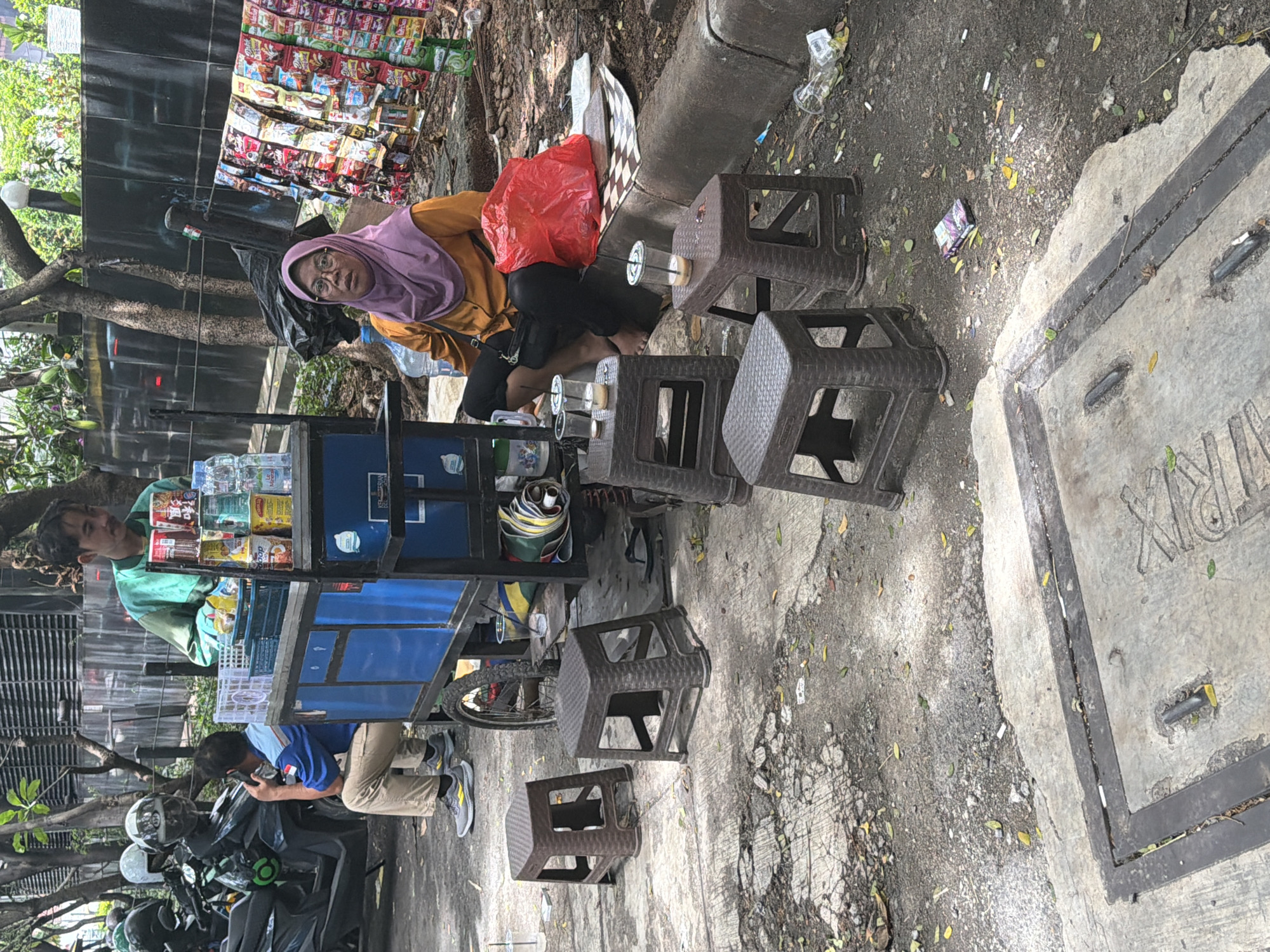 The scene is an outdoor vending operation in South Jakarta, Indonesia. A woman wearing a pink hijab and glasses is seated behind a food vending cart, with a red plastic bag next to her. Several brown plastic stools are arranged near her. The cart displays snacks and drinks. Cigarette butts and litter are scattered on the ground. Two individuals are near the cart: one wearing a green shirt is standing and one wearing a blue shirt is sitting. Several motorcycles are parked nearby. A concrete surface with metal bordering is visible on the right side of the frame.