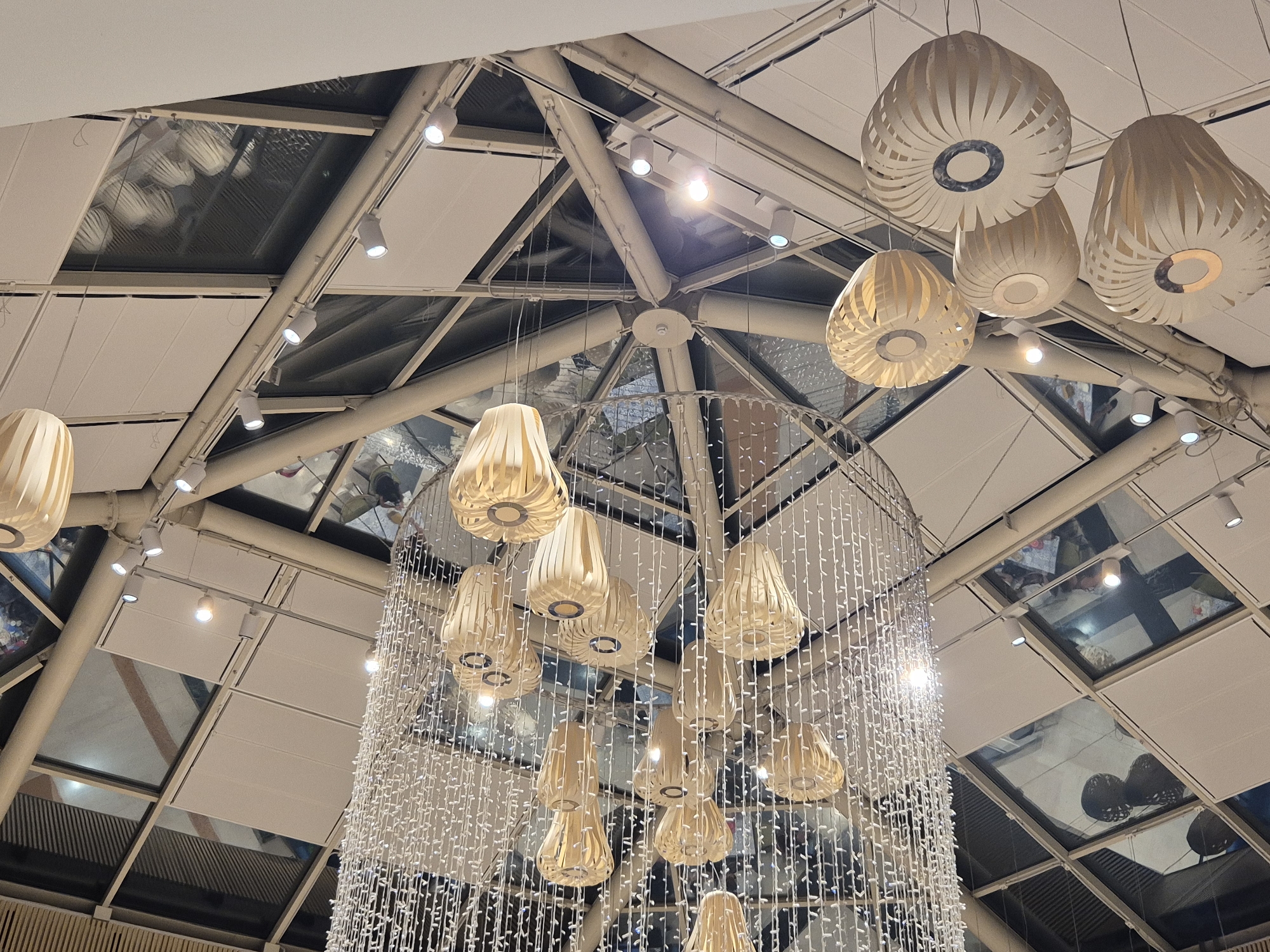 The image shows an indoor atrium with a complex, multi-level ceiling structure composed of white beams and panels, interspersed with reflective glass panels. Several decorative light fixtures are visible. Towards the top and right, there are several pendant lights with cream-colored, intricately woven shades, resembling beehives or seed pods. In the center and foreground, a large cylindrical structure is draped with a dense curtain of small, white LED lights, creating a shimmering effect. Hanging within this light curtain are more of the cream-colored pendant lights. The reflective glass ceiling panels reveal distorted reflections of the lights, ceiling structure, and possibly people or other objects below. There are also several white spotlights mounted on tracks along the ceiling beams, illuminating the space. The overall impression is of a modern, well-lit interior space, likely a shopping mall or public building. The reference to Taranto, Italy, suggests this is an indoor location within that city.