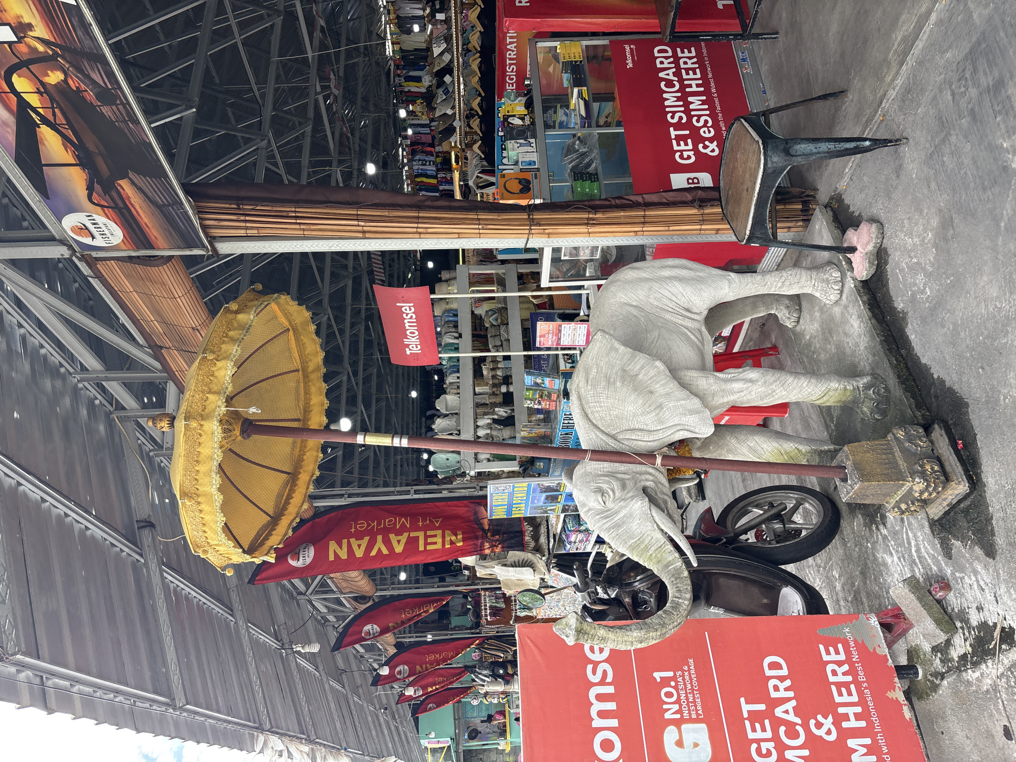 The image captures a scene at Nelayan Art Market in North Kuta, Indonesia. A grey stone sculpture of an elephant rests on the ground next to a red Telkomsel promotional sign, a small motorbike, and a golden ceremonial umbrella. A red banner with the words "NELAYAN Art Market" hangs above. There are shops and stalls visible in the background.