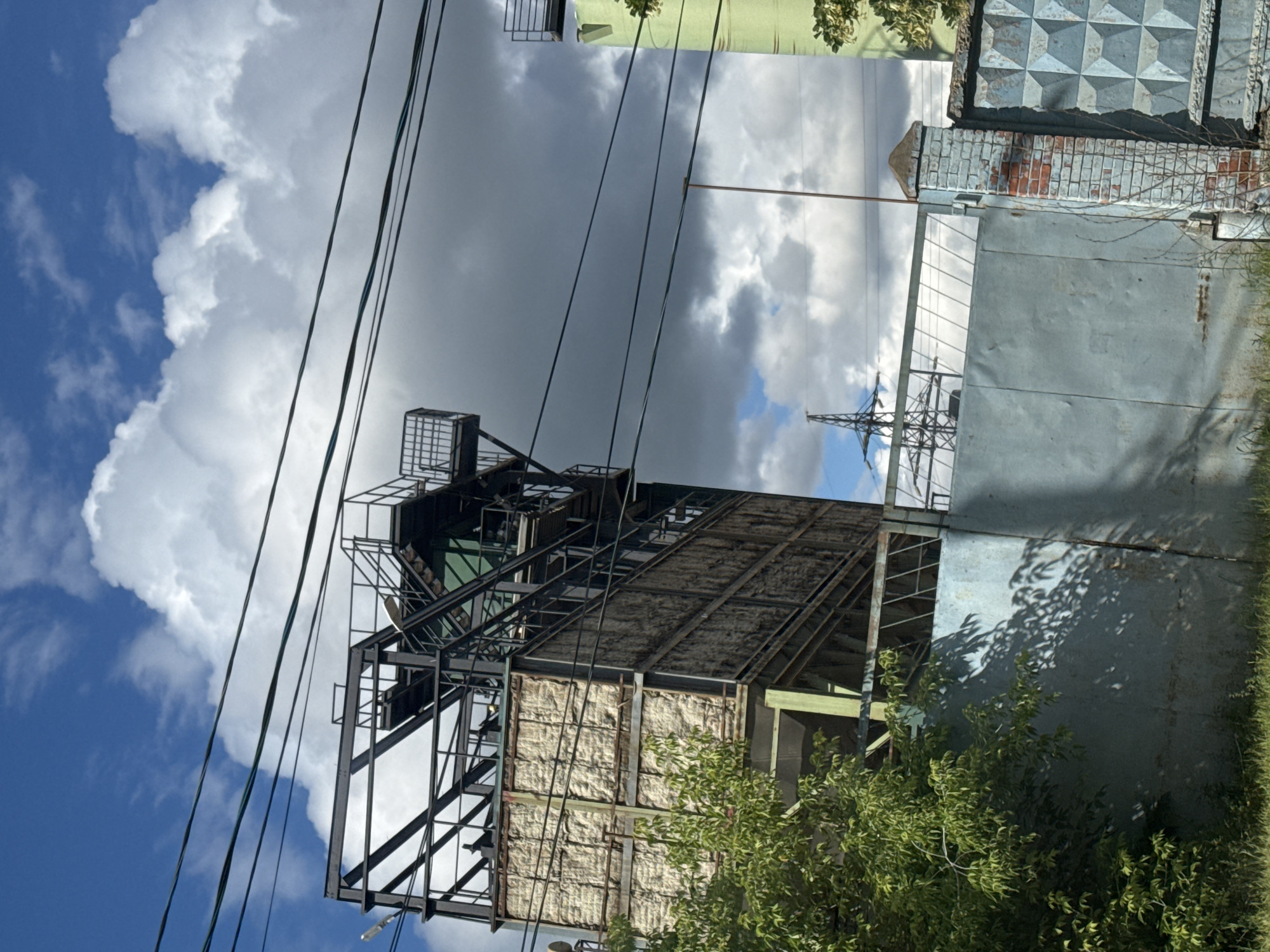 The image shows an industrial building with a metal frame, partially covered with insulation material. There is a large blue metal gate in the foreground, with a brick pillar to its right. A tree with green foliage is to the left of the gate, casting shadows. In the background, there are power lines, a transmission tower, and a green building. The sky is partly cloudy with blue sky visible. The scene appears to be in an industrial or semi-industrial area. The image is taken in the city of Khimki, Russia.