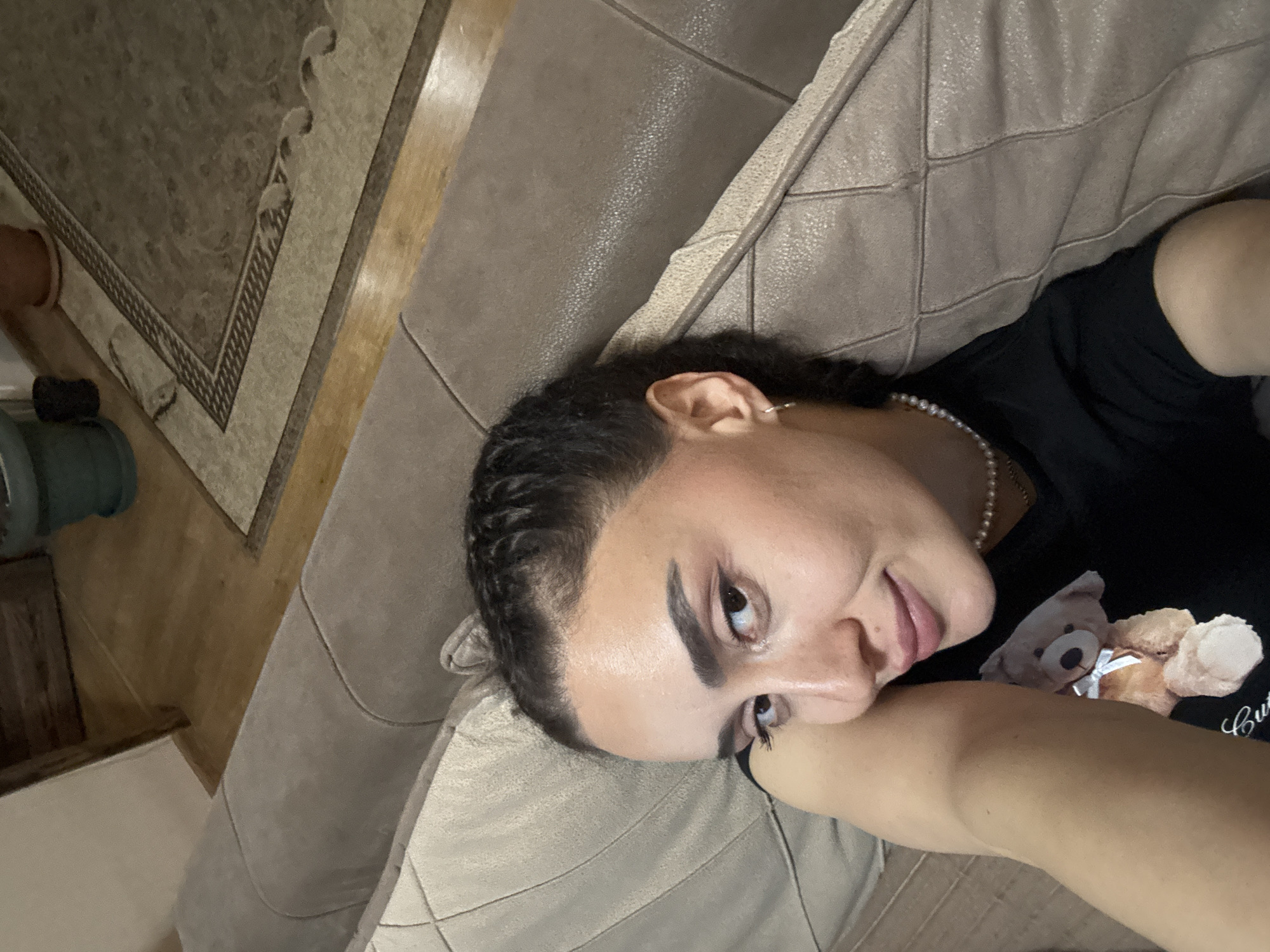 A person is lying on a beige quilted couch, wearing a black t-shirt with a teddy bear graphic and a pearl necklace. The individual has dark hair styled in tight braids and is looking at the camera. The setting appears to be indoors. Details to help ascertain the exact location in Tbilisi, Georgia are not visible in the image.