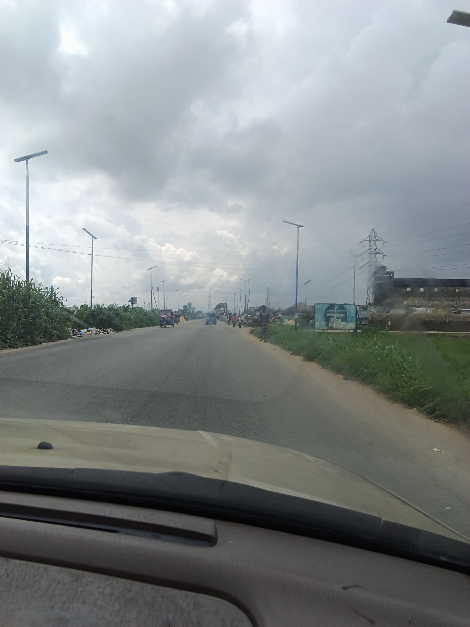 The image depicts a road scene in Yenegoa, Nigeria, under a cloudy sky. The road appears to be a multi-lane highway, with traffic visible in the distance. On the left side of the road, there is a dense growth of tall green vegetation, with some scattered refuse. Several modern streetlights with solar panels are installed along the roadside, suggesting this is a developed area.
Further down the road, there are several vehicles, including what appears to be a small bus or van and some smaller cars. A few people are visible walking along the side of the road or near parked vehicles. On the right side of the road, there are more structures, including what looks like a factory or industrial building with a tall transmission tower and power lines nearby. A blue sign with some white text is also visible on this side. The overall impression is of a busy but somewhat unkempt urban environment. The lighting suggests it could be daytime, but the heavy cloud cover gives the scene a somewhat subdued atmosphere. The presence of the transmission tower and the industrial buildings hint at an area with significant infrastructure.
