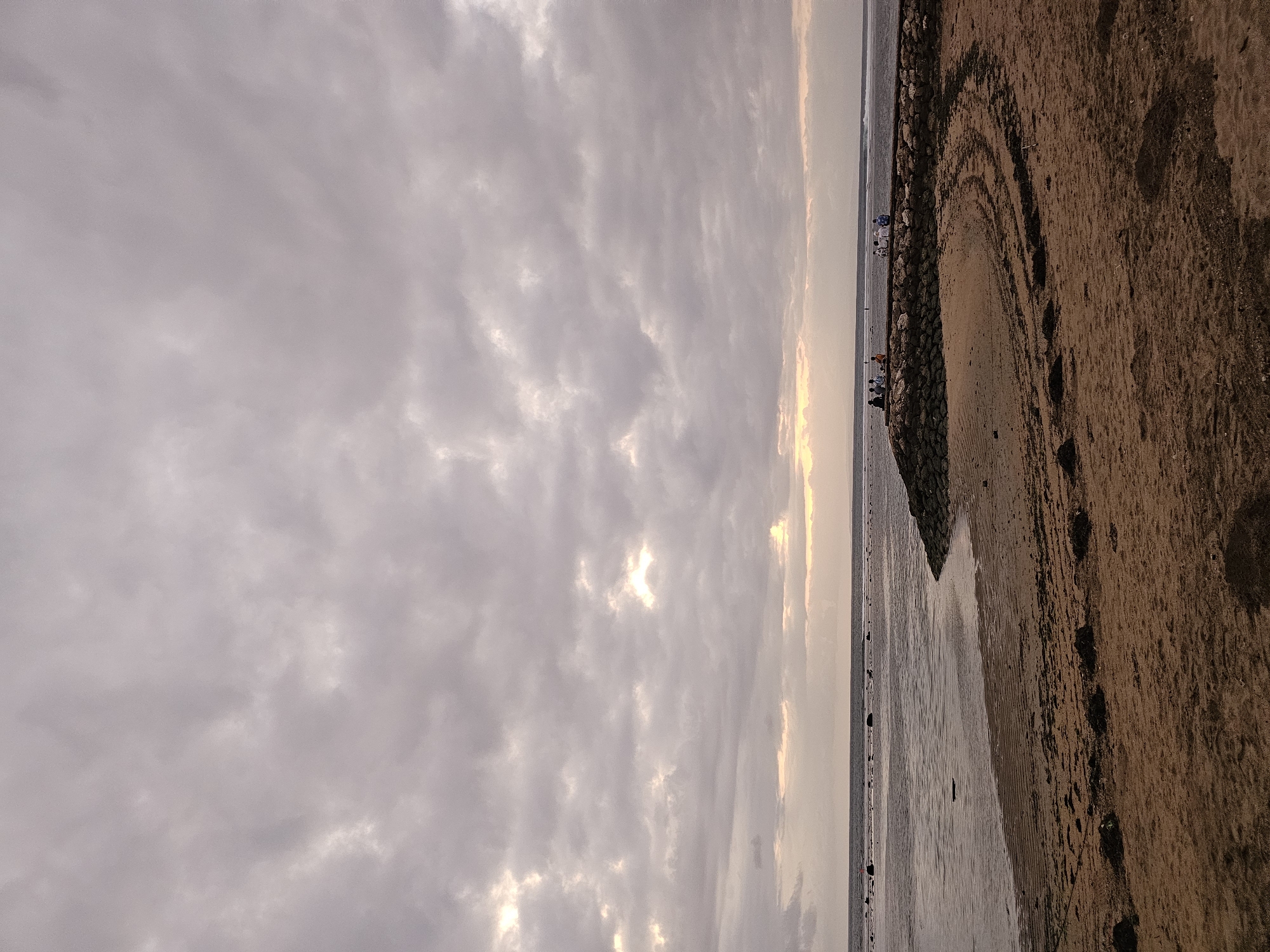 The image depicts a beach scene in Denpasar, Indonesia, under a cloudy sky. The foreground is dominated by sandy beach with scattered dark seaweed. A curved breakwater made of rough stones extends into the ocean, with several small groups of people sitting on top of it. The ocean is calm with a gentle surf visible on the right horizon. The sky is heavily overcast with dramatic cloud formations and patches of light breaking through. The overall atmosphere is serene and overcast, characteristic of a beach during or just before a storm.