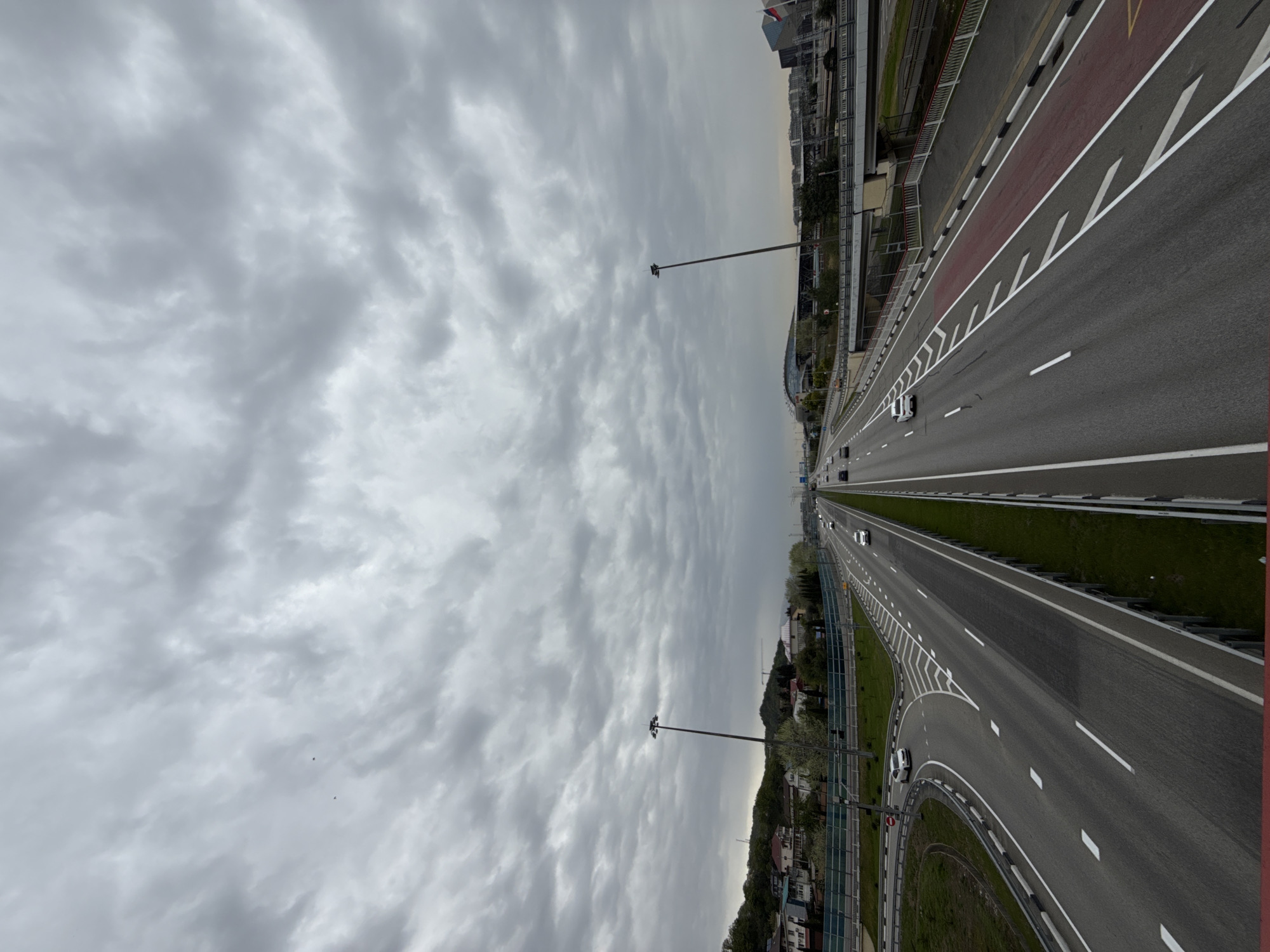 The image shows a multi-lane highway in Sirius, Russia, under a cloudy sky. The roadway has directional markings and painted areas on either side. There are street lights along the highway. Several cars are visible traveling in both directions. Buildings and greenery are seen on the sides of the road.