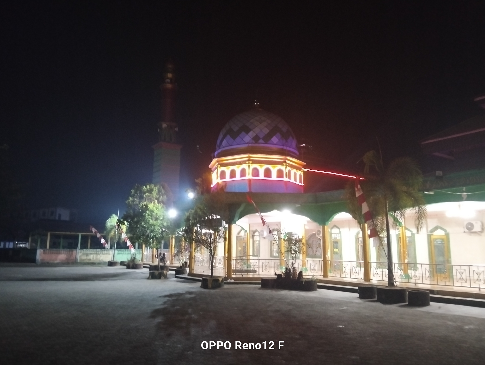 The image shows a mosque at night in Buntok, Indonesia. The mosque is lit up with colorful lights, and there is a tall minaret. In the foreground, there is a paved area with some trees and flags. The sky is dark, and there are no people visible in the image. The overall scene is tranquil and serene.