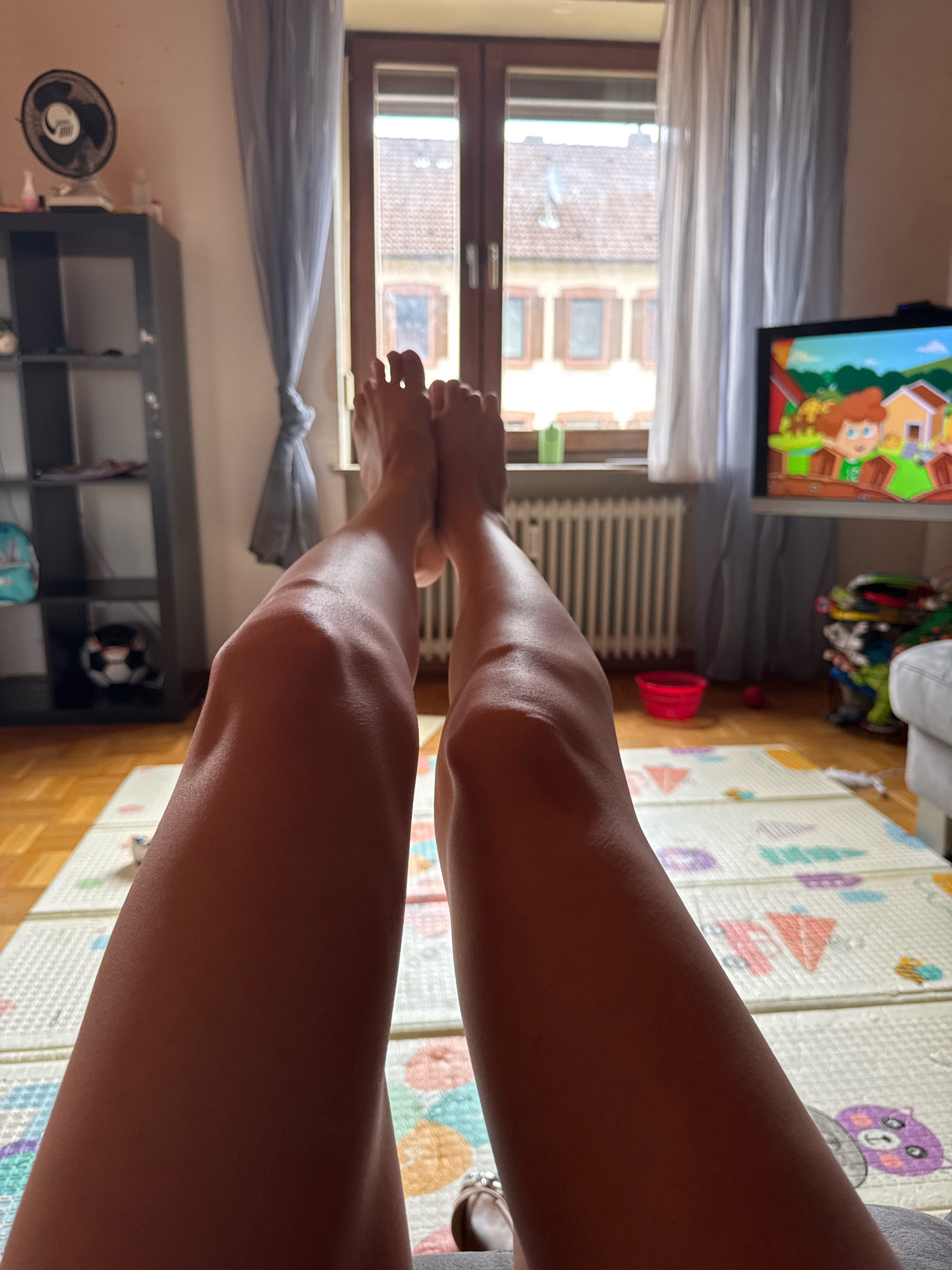 The image shows a close-up view of a person's legs from the knees down, with their feet positioned in the foreground. The person is lying down, as indicated by their relaxed posture and the way their legs are extended. They are indoors, in what appears to be a living room or play area.
In the background, a window overlooks buildings with red tile roofs, typical of European architecture. The window is draped with light blue curtains. To the right of the window, a television screen displays a colorful cartoon with animated characters. A radiator is visible beneath the window.
To the left of the window, a tall gray bookshelf contains various items, including a black fan on top and a soccer ball on a lower shelf. The floor is made of light-colored wood, and a large, padded play mat with colorful geometric patterns and cartoon animals is spread out. A red bucket and a small red ball are on the floor.
The lighting suggests it is daytime, with natural light coming from the window. The image has a casual, domestic feel, possibly capturing a moment of relaxation or entertainment. The presence of the play mat and cartoon on TV suggests the presence of children, though they are not visible in the frame. The overall setting appears to be a residential space. The specific location in Überherrn, Germany, is not directly identifiable from the visual cues alone, but the architectural style and general environment are consistent with a European setting.