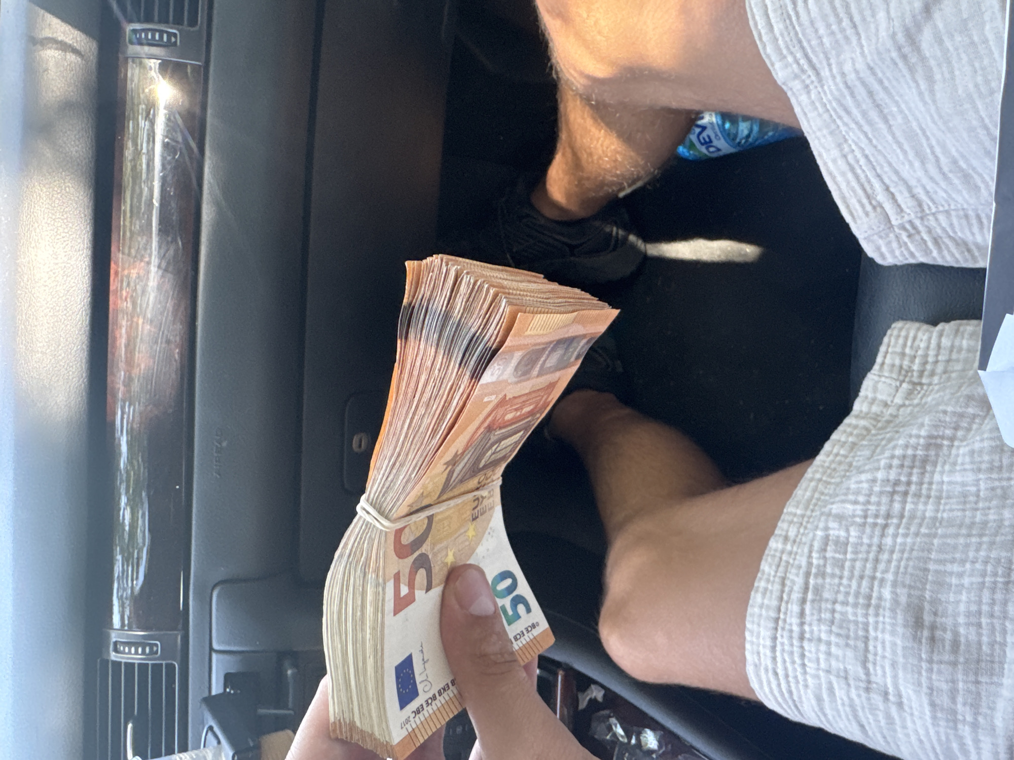 The image depicts a close-up view from inside a car, focusing on a person's hand holding a large stack of 50 Euro banknotes. The money is secured with a white rubber band. The background shows the car's interior dashboard, including air vents and wood-grain trim. The lower part of the image reveals the person's legs, clad in light-colored, textured shorts, and a black sneaker. A blue bottle of "DEV" water is visible in the car's cup holder. The scene suggests the individual is a passenger or the driver of the vehicle. The lighting indicates it is daytime, with sunlight illuminating parts of the interior. No specific location within Sofia, Bulgaria, can be determined from the visual information provided.