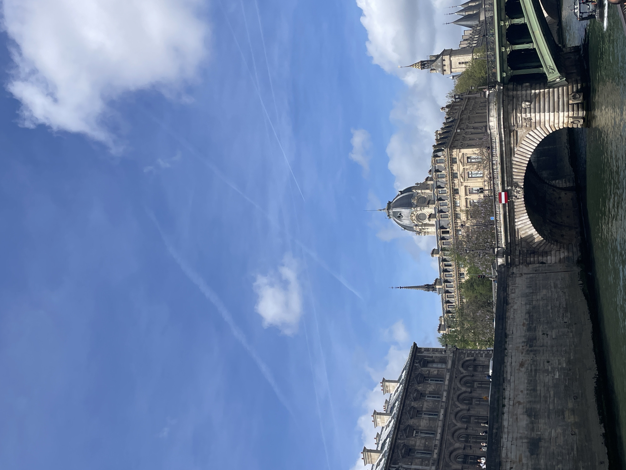 The image captures a view of Paris, France, specifically along the Seine River. On the right, an arched bridge structure is visible, possibly Pont Neuf, with visible detail of the stone construction and the flowing river beneath. Adjacent to the bridge, a stone embankment or quay lines the river. The architectural style of the buildings lining the river suggests traditional Parisian design with visible windows, roofs, and stonework. The sky above is blue with scattered clouds and vapor trails from airplanes. The overall scene suggests a clear, sunny day in a historical part of Paris, showcasing architectural elements, river views, and cityscape.