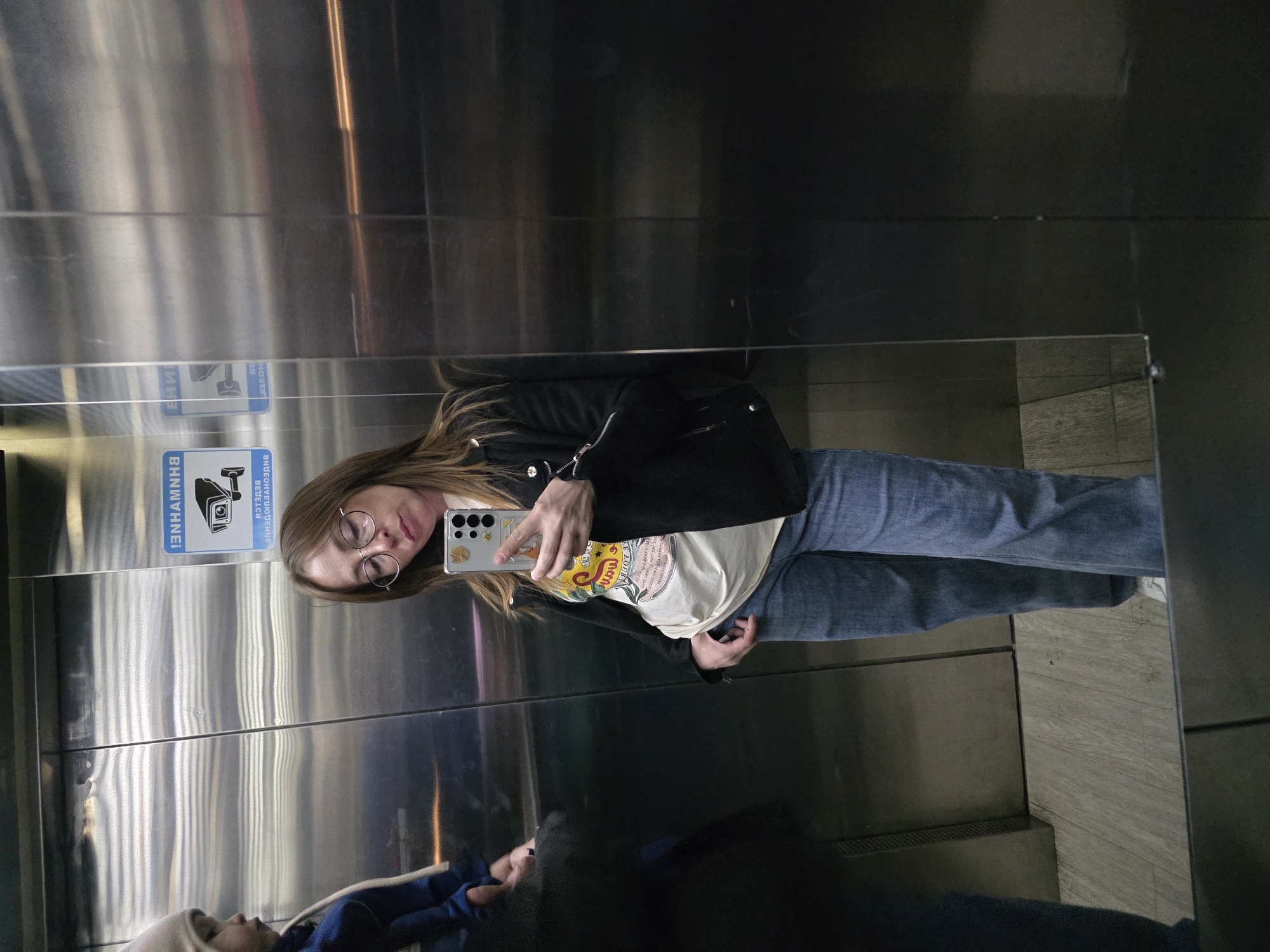 The scene depicts the interior of an elevator located in the city of Votkinsk, Russia. The elevator features brushed metal walls, one of which acts as a reflective surface showing the scene as a mirror selfie. The floor is covered with a light-colored, wood-grain laminate.
A female adult, approximately 30-40 years old, is the central subject, capturing the image via a mirror selfie using a light-colored smartphone encased in a transparent, possibly cat-themed, case. She has shoulder-length brown hair and wears round eyeglasses. Her attire consists of a black leather-style jacket with metallic zippers and snaps, a light-colored t-shirt with a yellow and red graphic design, and blue wide-leg jeans. Her left hand holds the phone, while her right hand is positioned near her right hip.
To the left of the female adult, a child is partially visible, wearing a light-colored beanie and a blue long-sleeved garment. Only the child's head and upper torso are within the frame, with the child looking towards the left side of the elevator, away from the camera.
A blue and white sticker is affixed to the metal wall, displaying a CCTV camera icon and the mirrored Russian text "ВНИМАНИЕ! ВЕДЕТСЯ ВИДЕОНАБЛЮДЕНИЕ" (ATTENTION! VIDEO SURVEILLANCE IS IN PROGRESS).