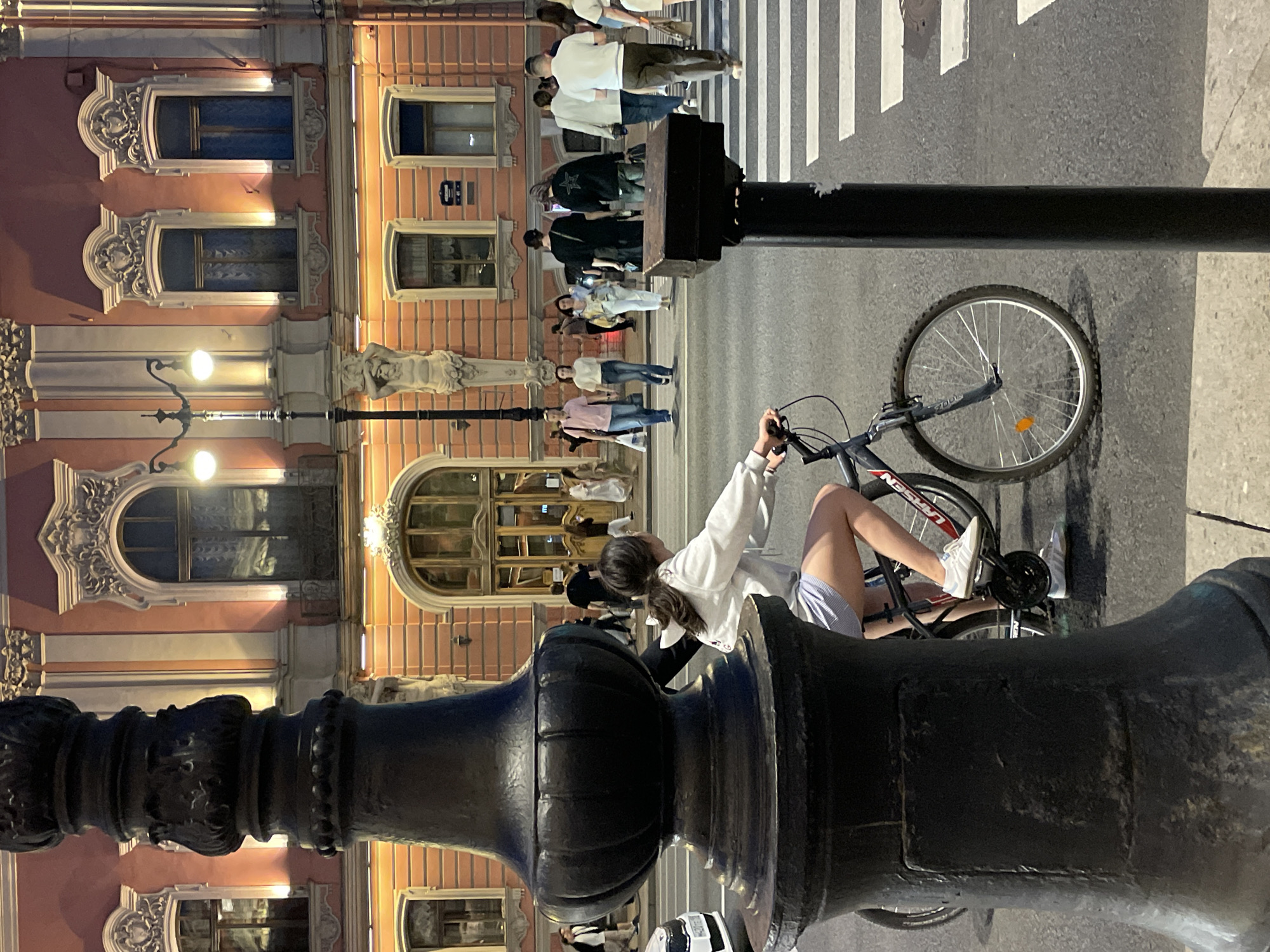 The image depicts an evening street scene in Saint Petersburg, Russia, specifically at Liteyny Prospekt, 57. The prominent structure in the background is a multi-story building, distinctive for its coral-pink facade adorned with elaborate white stucco architectural detailing, including ornate window surrounds, cornices, and a grand arched entrance at the ground level. A white caryatid sculpture supports an upper element to the left of the main entrance. The building is illuminated by warm artificial lighting, casting reflections in its windows.
In the immediate foreground, to the left, a large, dark, ornate cast-iron lamppost occupies a significant portion of the frame. In the mid-ground, a young female, with long dark hair, wearing a white long-sleeved top and light blue and white striped shorts, is seated on a dark-colored bicycle. She is wearing white athletic shoes and her left foot is positioned on a pedal. Her hands rest on the handlebars, and she looks towards her right (viewer's left). The bicycle has an orange reflector on its front wheel and "LARSEN" visible on its frame.
Behind the cyclist, a dark asphalt road features a white-striped pedestrian crosswalk. Numerous pedestrians are visible crossing the street or walking on the sidewalk. Individuals include a man in a pink polo shirt and jeans, a woman in a white t-shirt and jeans, a man in a black long-sleeved shirt with a white star on the back, and a man in a white polo shirt and olive pants. On the far left, partially obscured by the lamppost, is the front section of a white vehicle, likely a Skoda. A slender dark pole, presumably for a traffic or pedestrian signal, stands to the right of the crosswalk. The overall environment is an urban setting under artificial illumination, indicating either twilight or nighttime.