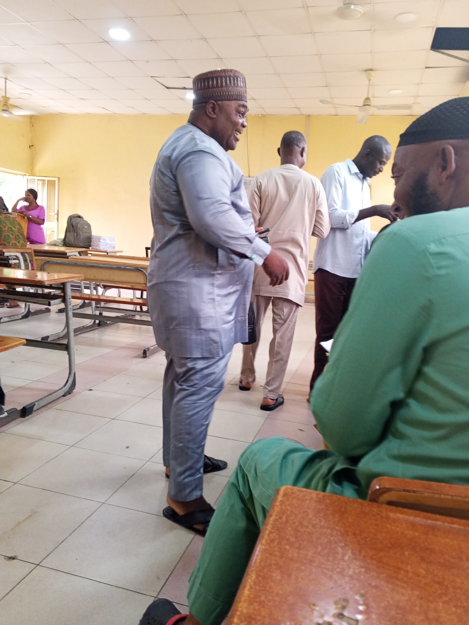 In Lokoja, Nigeria, a group of men dressed in traditional attire are gathered indoors, possibly in a classroom or meeting hall. The room has a tiled floor and a yellow-painted wall. Several desks with benches are visible, some with textbooks stacked on them. In the background, a woman in a pink outfit stands near a doorway. The men are engaged in conversation, with one man in a light grey outfit and cap in the foreground smiling. Another man in a green shirt and cap is seated at a desk, facing away from the camera. The lighting appears to be from overhead fluorescent lights.