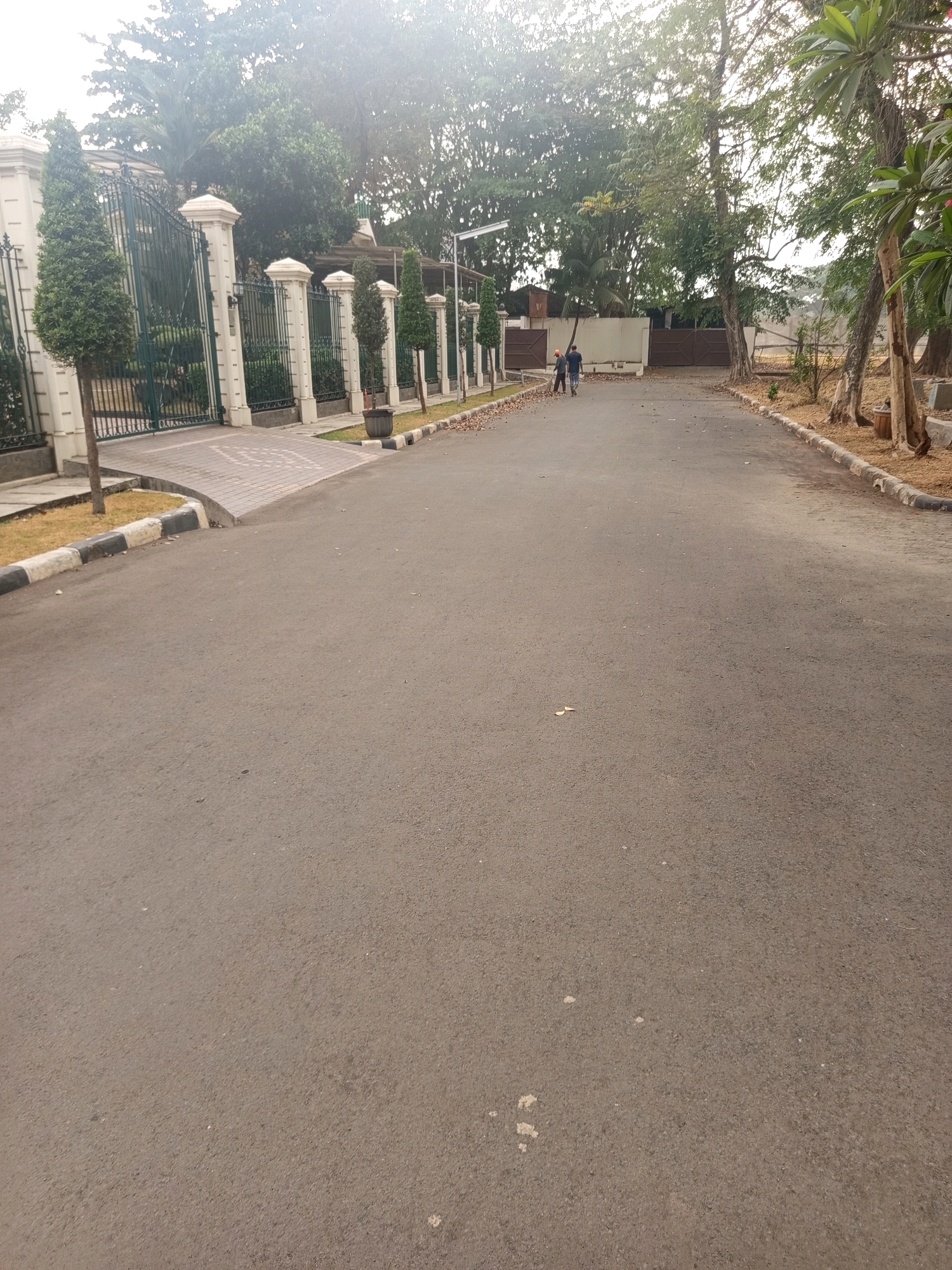 The image shows a paved road leading towards a large gated property. The gate is green and ornate, supported by white pillars. There are manicured trees and hedges lining the road and the property entrance. In the distance, two individuals are walking away from the camera down the road. They are wearing casual clothing. The environment appears to be a residential or commercial area with mature trees and some landscaping. The road is asphalt and appears well-maintained. This scene is located in the city of West Jakarta, Indonesia, specifically in the Permata Hijau neighborhood, known for its upscale residences and lush greenery.