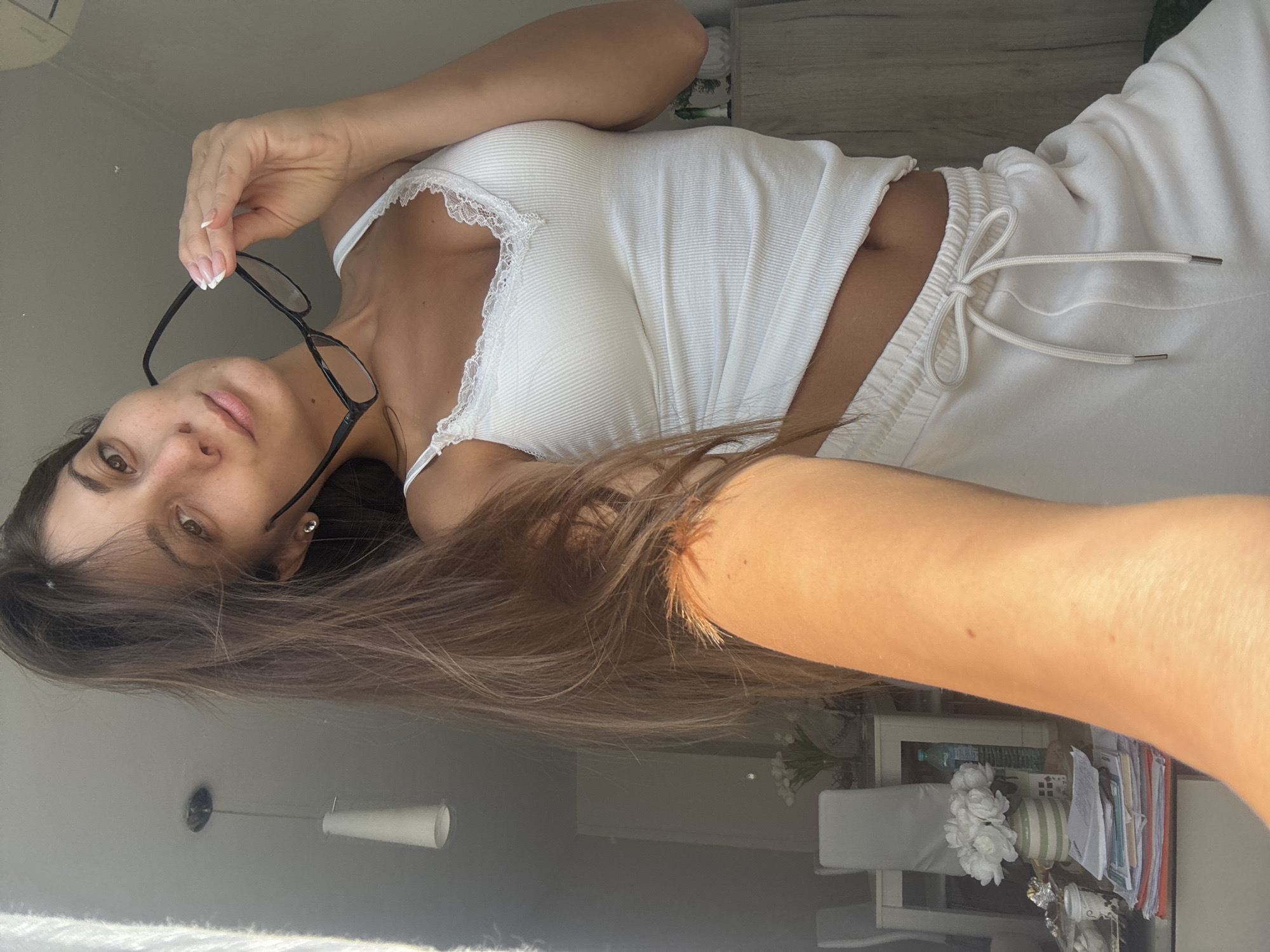 A woman with long brown hair is holding black framed glasses. She is wearing a white ribbed tank top with lace trim and white drawstring sweatpants. The background appears to be an interior space with a light fixture and furniture. There is no indication of the location being Burgas, Bulgaria in the image.