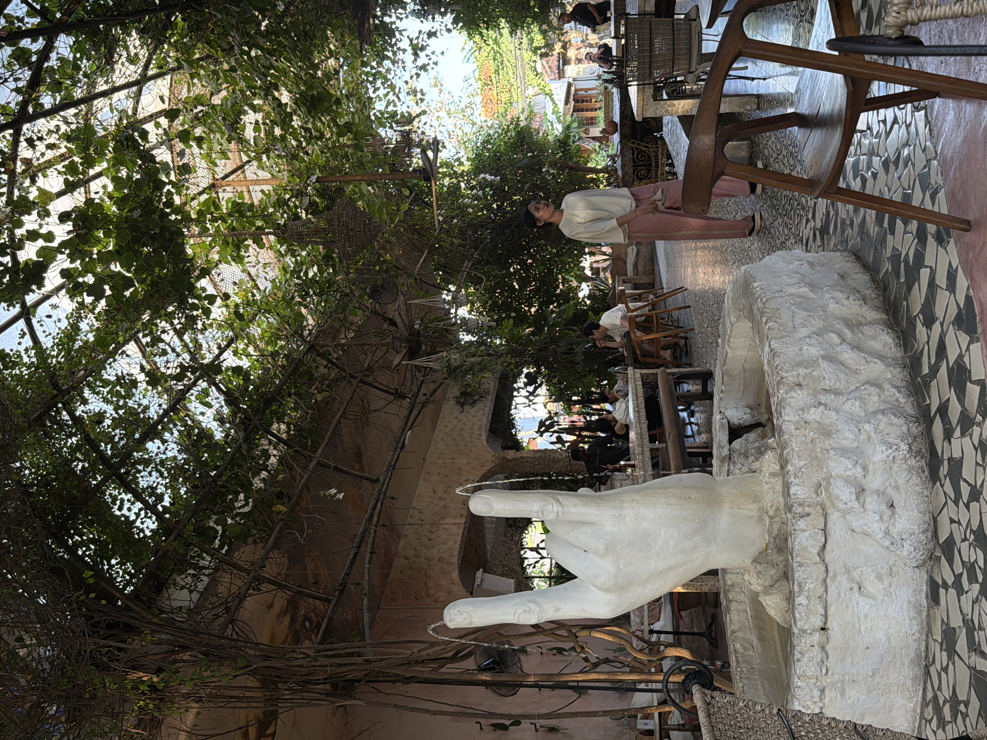 The image depicts an outdoor cafe in North Kuta, Indonesia. A white sculpture of a hand making a peace sign with water spraying from the index and middle fingers serves as a fountain in the foreground. The cafe has a rustic ambiance with a roof covered in lush green vines and trees, creating a natural canopy. Seating areas with wooden chairs and tables are visible throughout the space, interspersed with plants and ambient lighting. In the middle ground, a woman in a white top and pink pants walks across the mosaic-tiled floor. Several other patrons are seated at tables in the background, engaged in conversation or dining. The overall scene suggests a relaxed and vibrant atmosphere in a tropical setting.