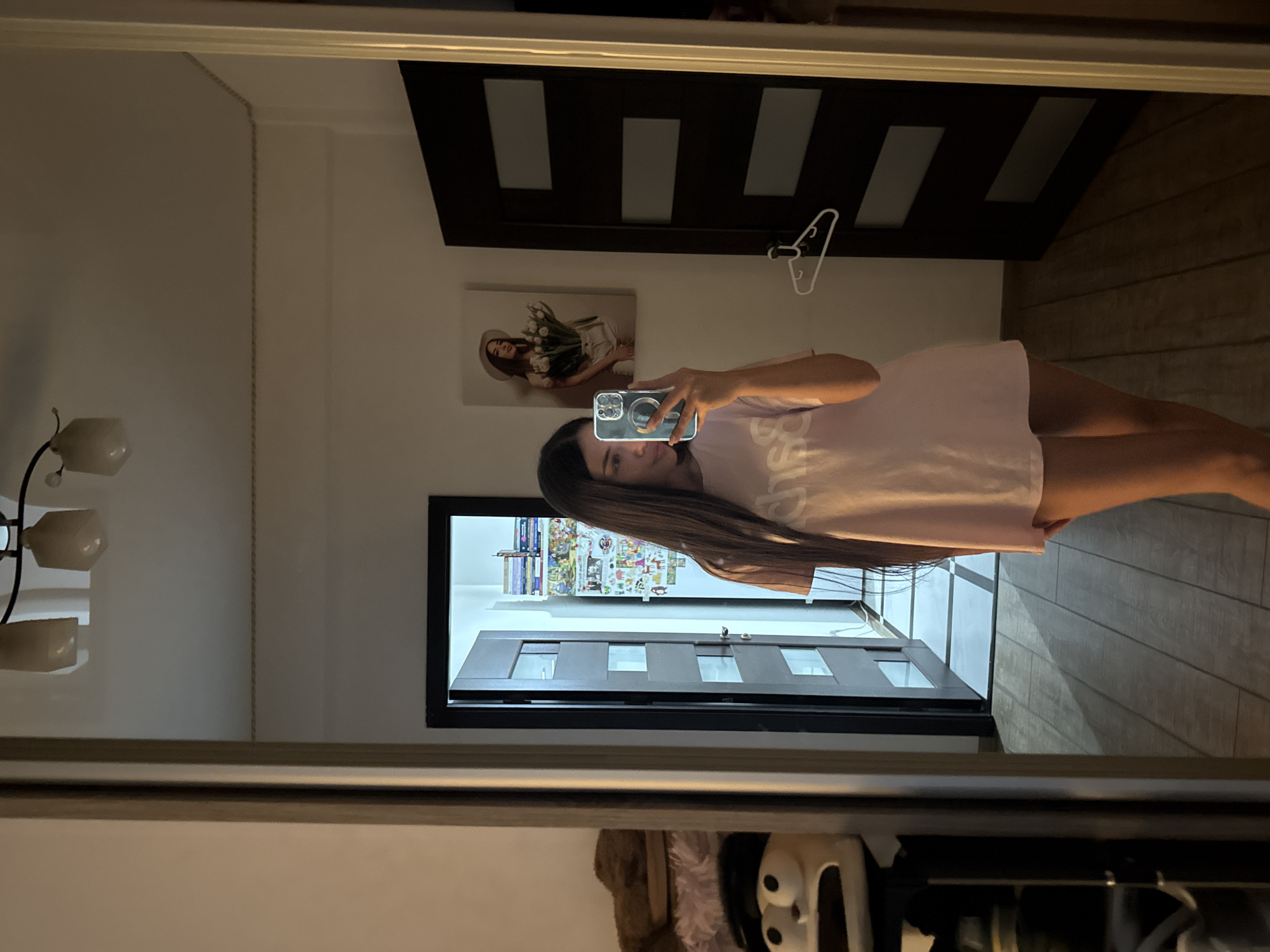 A young woman with long, dark hair is taking a mirror selfie in what appears to be a bedroom or dressing area. She is wearing a light pink t-shirt and her legs are visible. The room has wooden flooring and several doors, one of which is open, revealing a hallway with posters on the wall. A large mirror dominates the foreground, reflecting the woman and the room. To the left, there is a shelf with stuffed animals. A framed picture of a woman holding flowers hangs on the wall behind the woman in the mirror. The lighting suggests it is evening or the room is dimly lit. There is no specific indication of the location being in Chișinău, Moldova.