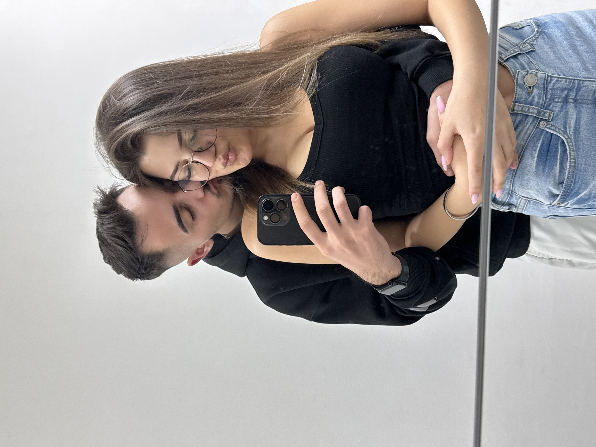 A photograph depicts a young couple in a mirror selfie. The woman, with long brown hair and eyeglasses, wears a black top and jeans. The man wears a black hoodie and holds the smartphone, capturing the image. Their faces are close together, as if posing for a kiss. The scene is likely indoors, given the plain white wall and the presence of the mirror. There is no indication of Sofia, Bulgaria or any outdoor location.