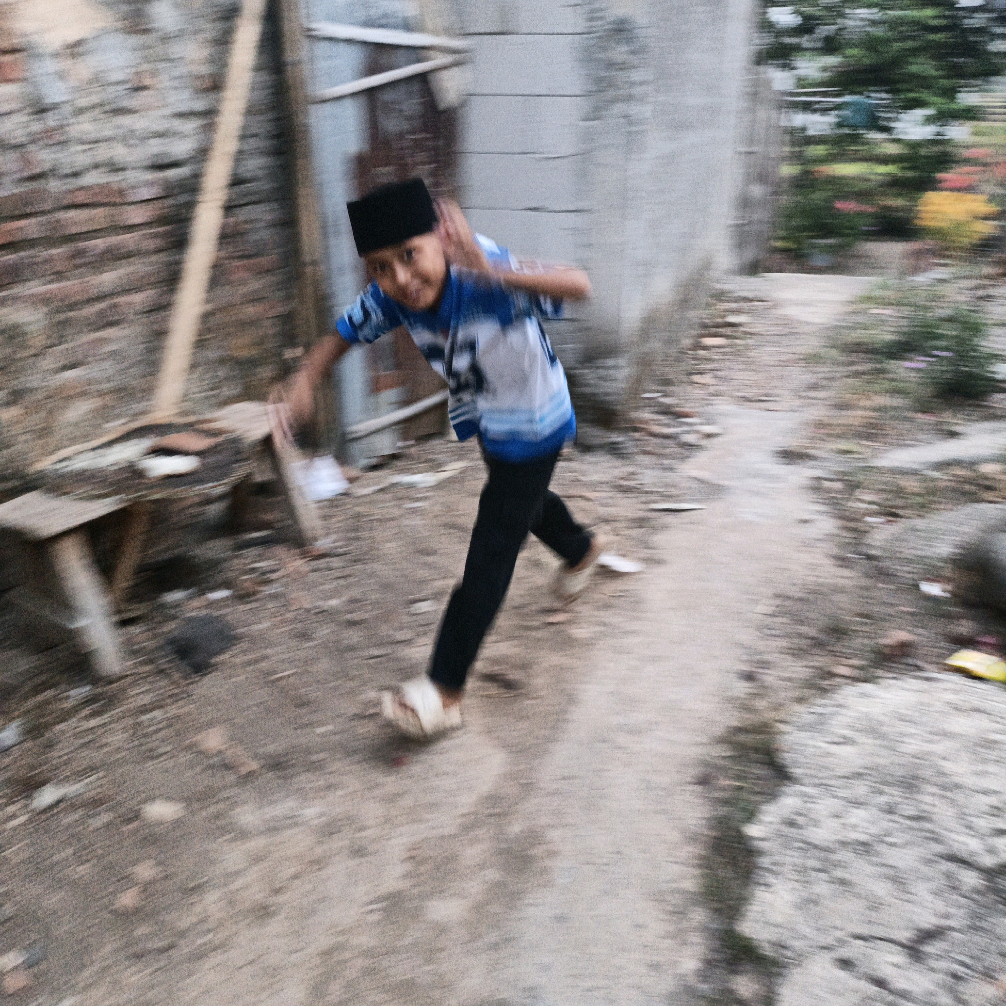 A young boy in a black peci and a blue and white patterned shirt is captured in motion, running or walking quickly on a dirt path. He is wearing black pants and light-colored sandals. His right arm is raised and bent, with his hand near his head. The background consists of a brick wall on the left, a light-colored building with horizontal lines on the right, and a garden with blurred foliage and flowers behind the building. A wooden bench is visible on the left side of the path. The image appears to have been taken in Tanahbaya, Indonesia, given the context of its origin. The motion blur suggests movement, possibly a child playing or running.