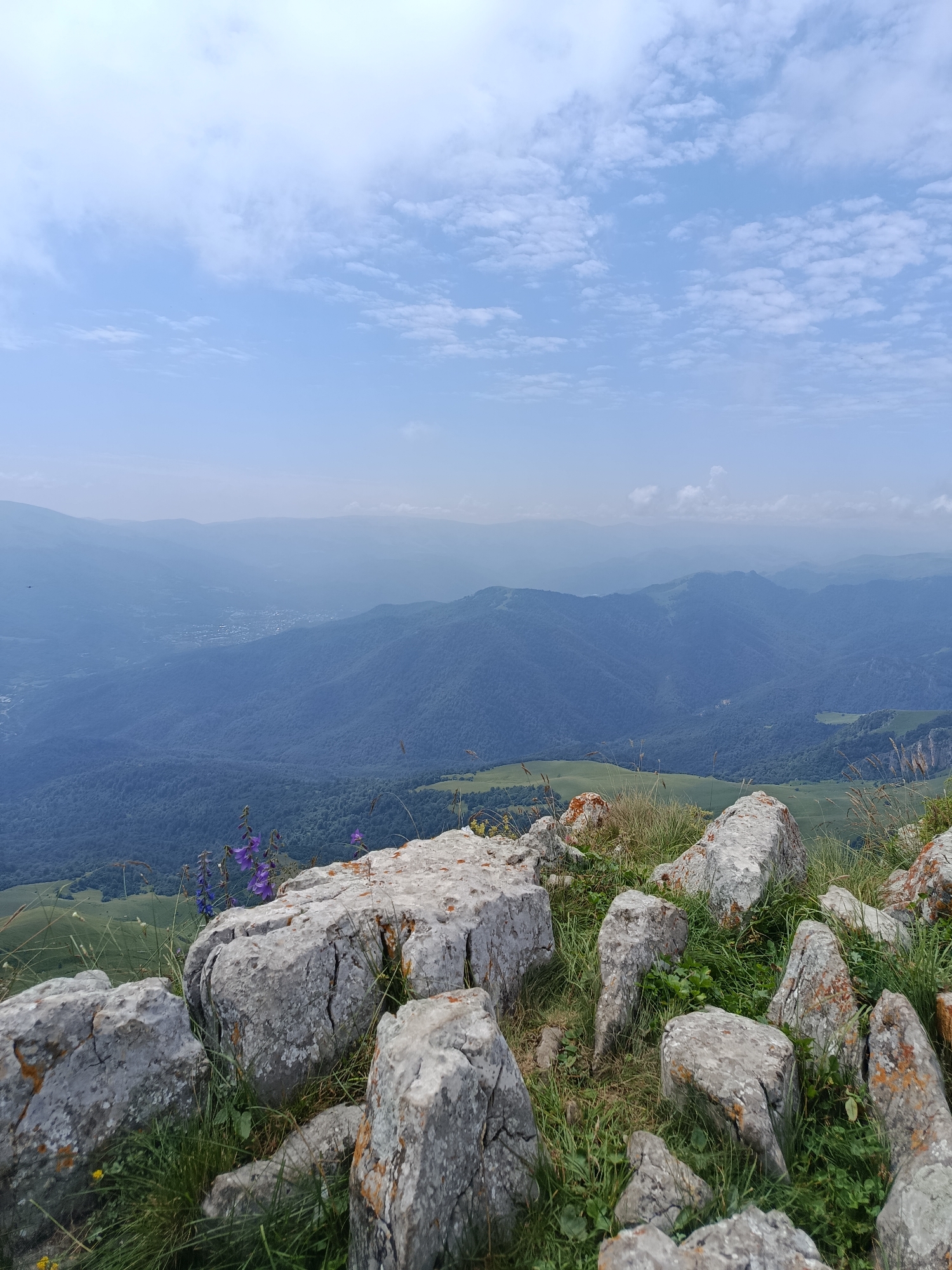This is a landscape photograph taken from a high vantage point, likely a mountain ridge, overlooking a valley. The foreground is dominated by large, grey rocks covered in patches of orange lichen and surrounded by green grass. A cluster of purple wildflowers is visible among the rocks in the mid-foreground.
In the background, a vast expanse of rolling hills and mountains recedes into the distance, fading into a hazy blue atmosphere. A small town or village can be seen nestled in the valley floor, with buildings appearing as small specks. The sky above is a light blue with scattered white clouds.
The overall scene depicts a natural, mountainous environment with signs of human habitation in the valley below. The photograph is taken in daylight, suggesting it is daytime. There are no people or animals visible in the image. The location is identified as Teghut, Armenia, a region known for its mountainous terrain and natural beauty.