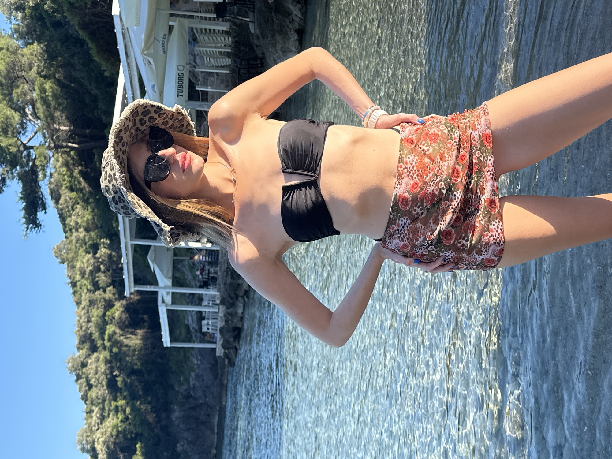A young adult female stands in clear, shallow water, likely a cove or beach, in Kubasi, Montenegro. She is positioned with hands on hips, body slightly angled right, and head turned left. She wears a black strapless bandeau bikini top, a sheer floral and leopard print mini-skirt, a leopard print bucket hat with a frayed brim, and dark sunglasses. A delicate gold-toned necklace and multiple light-colored bracelets on her right wrist are visible, along with blue nail polish on her fingers. The water's surface is rippled, revealing a rocky bottom. In the midground, white open-air structures with rectangular pillars, tables, and chairs line a rocky shore. A "TUBORG" sign is visible on a white umbrella or awning among these structures. Dense green Mediterranean vegetation covers the hills behind the structures. The sky is clear blue, indicating strong sunlight.