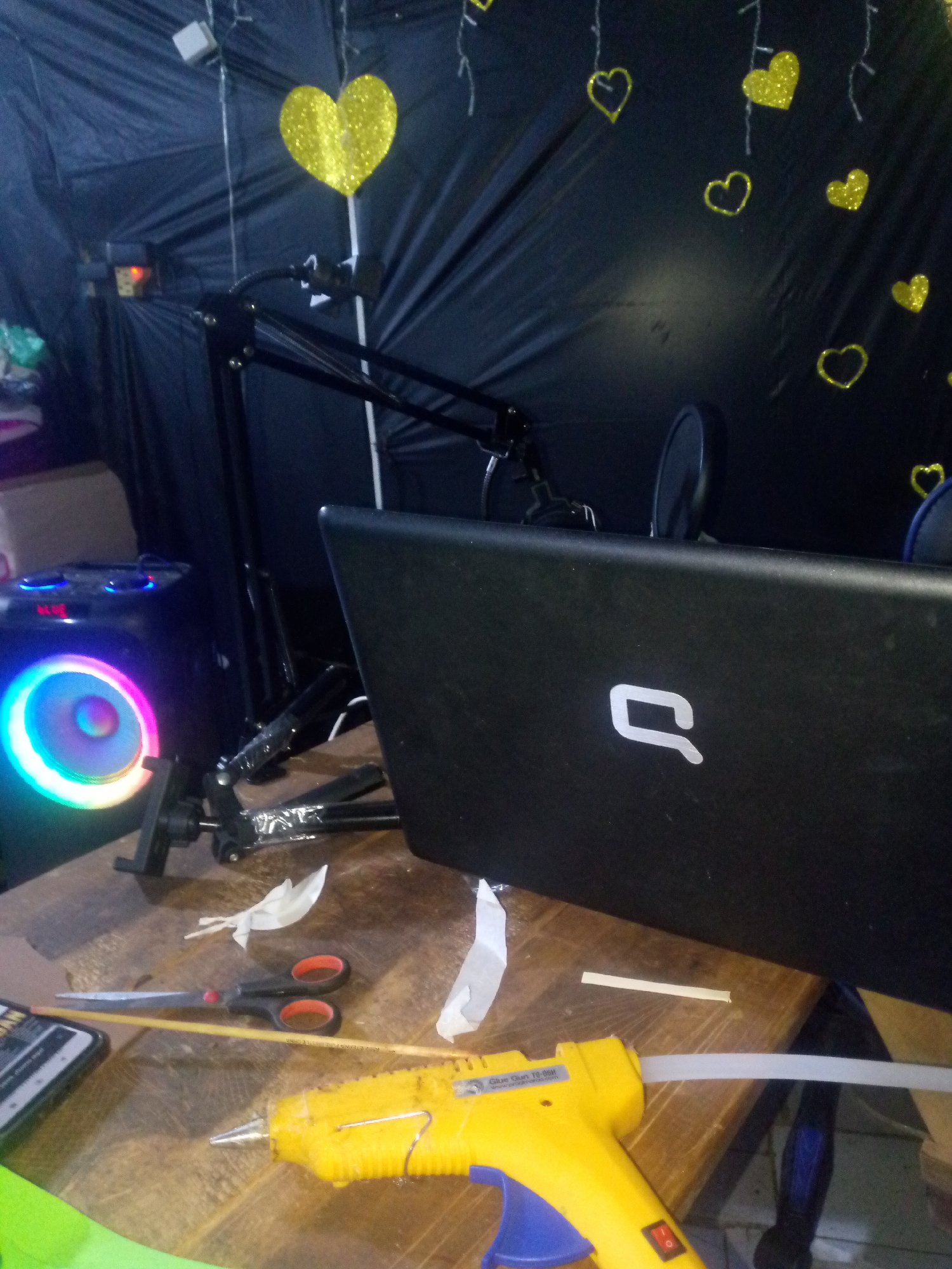The image shows a work desk with various items, including a yellow glue gun with a glue stick inserted, a pair of scissors with black and red handles, a computer monitor, and a black microphone stand. A circular object with rainbow-colored lights is also visible. The background features a black sheet decorated with yellow heart-shaped ornaments. The image does not provide any information about the location in the city of Juan Griego, Venezuela.