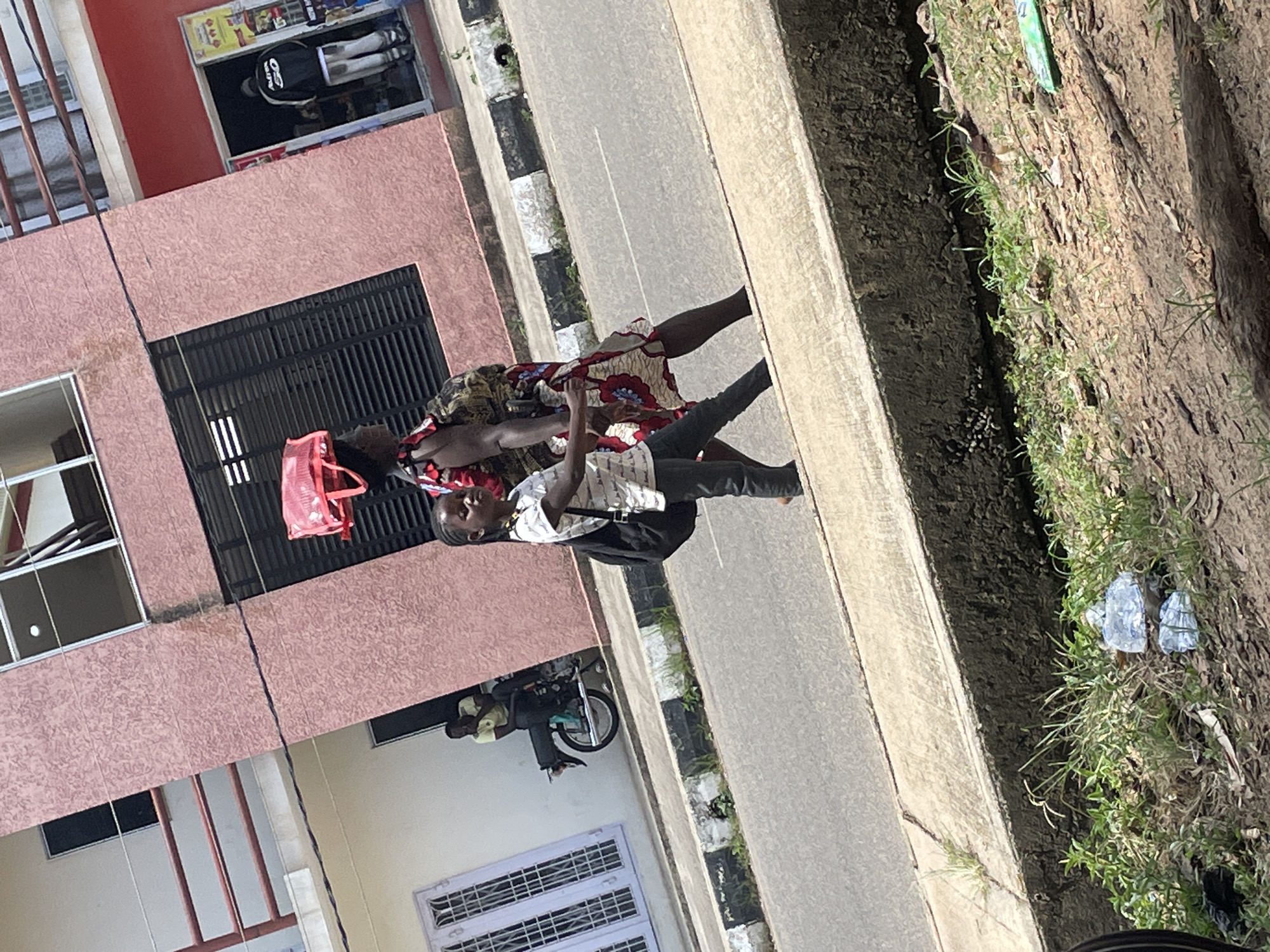In the city of Jos, Nigeria, a woman and a young girl walk along a paved road beside a pink textured building. The woman carries a red bag balanced on her head and wears a patterned dress. The girl, dressed in a white patterned shirt and dark pants, carries a black backpack. In the background, a man sits on a motorcycle near a doorway. The road has a black and white striped curb. Green grass and discarded plastic bottles are visible on the embankment next to the road.