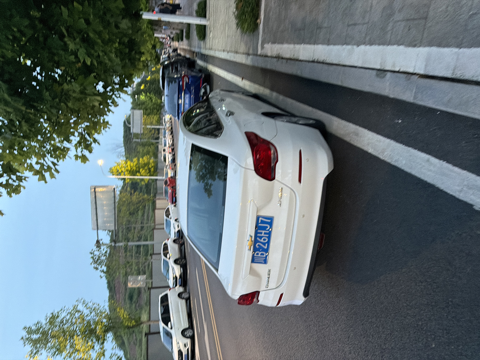 The image depicts a street scene in Jiangjin, China, likely during the late afternoon or early evening, given the soft lighting and clear sky. The foreground is dominated by the rear of a white Chevrolet sedan, identifiable by its emblem and license plate, which reads "B-26HJ7." The car is parked on the right side of the road, next to a sidewalk. Multiple other vehicles are visible parked and driving on the street, including a white minivan, a white SUV, a red sedan, and several other cars further down the road. The street is lined with trees, casting shadows on the pavement. In the background, a hill covered in vegetation is visible, with road signs and streetlights indicating an urban or semi-urban environment. The overall atmosphere is calm and ordinary, capturing a typical moment in city life.