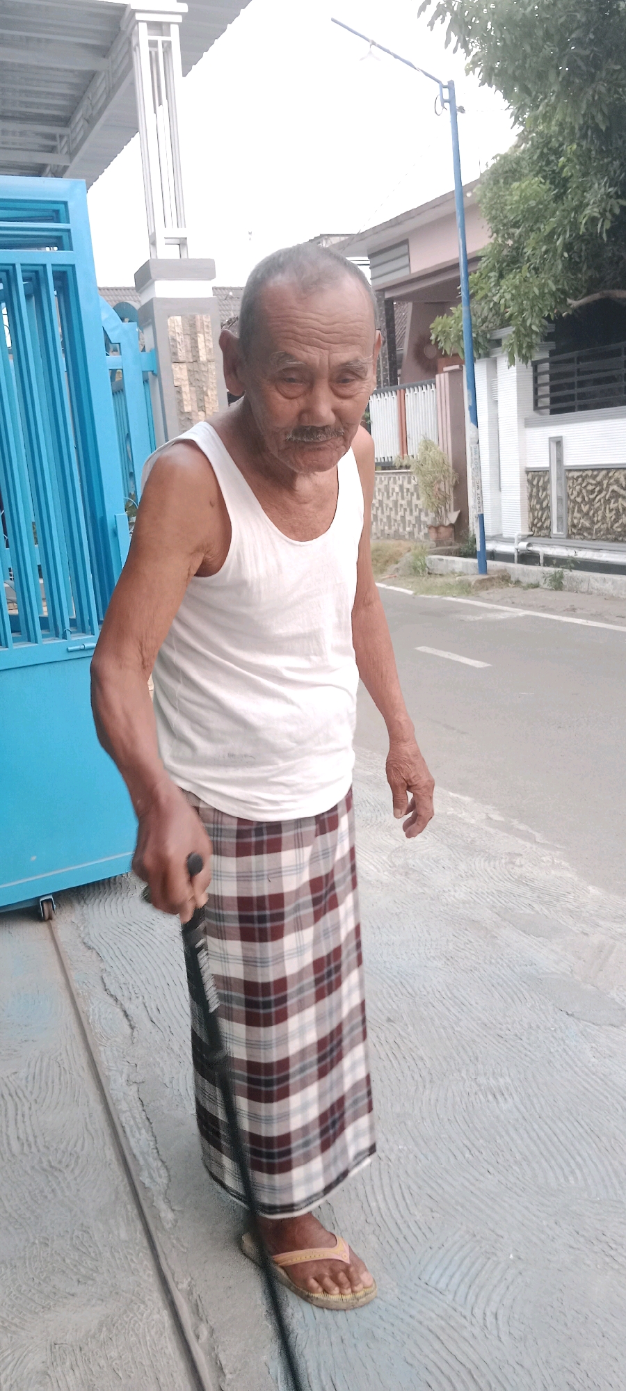 An elderly East Asian man stands in front of a blue metal fence and a paved walkway, in front of a street and some buildings. He is wearing a white tank top, a plaid sarong, and brown flip-flops. He is holding a black cane in his right hand, and is walking. The man has thinning grey hair, weathered skin, and a mustache. He appears to be walking towards the right of the frame. The scene takes place in Soko, Indonesia.