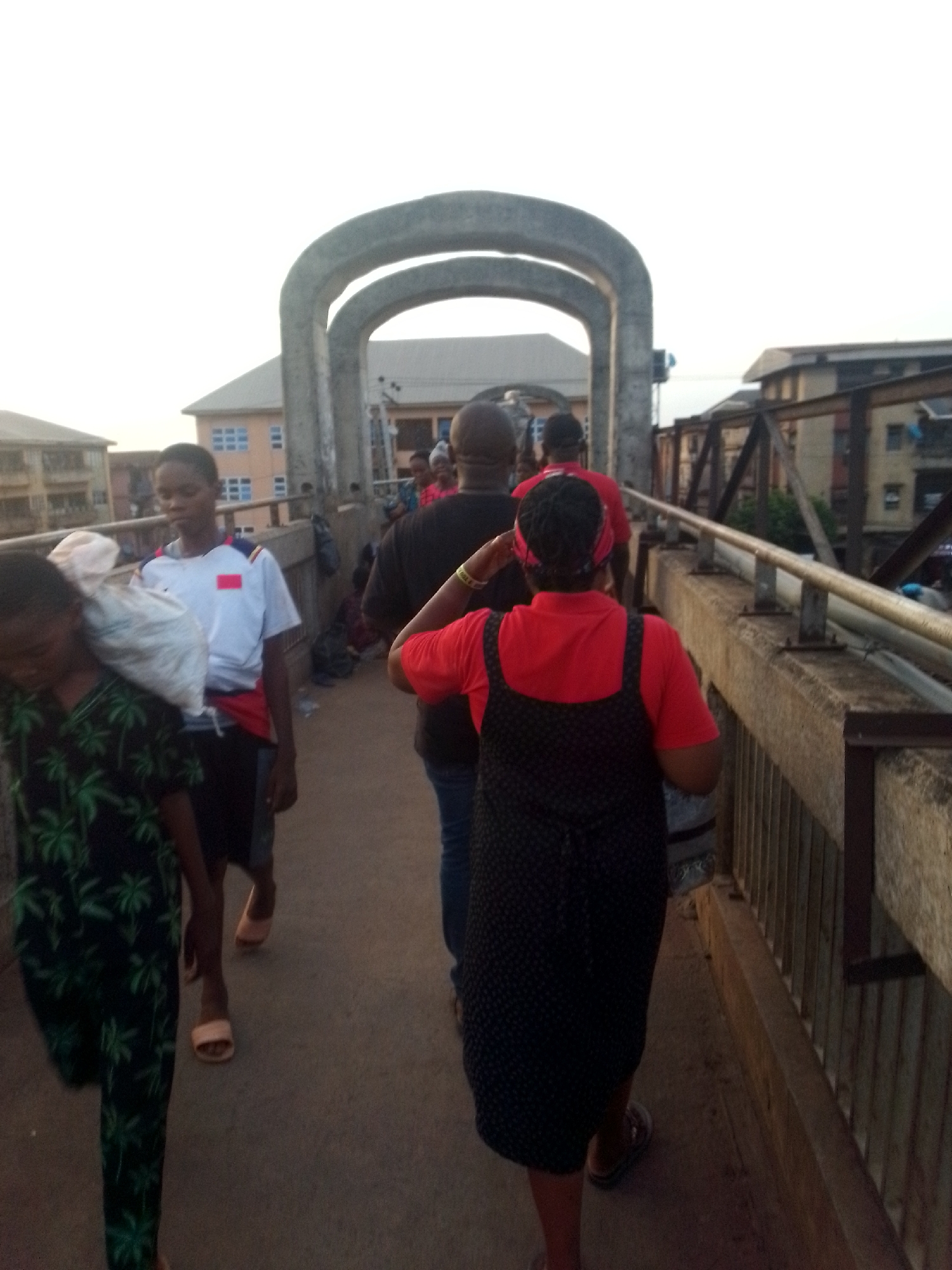 People walk across a concrete pedestrian bridge in a city. The bridge has three arched supports. Buildings are visible in the background. One person carries a bag over their shoulder. The people are moving away from the viewer.