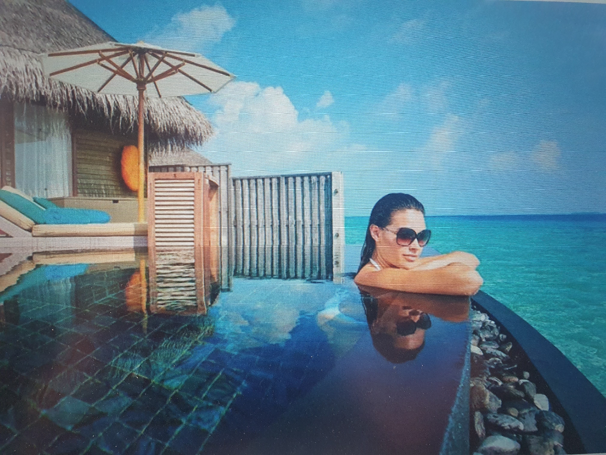 The image depicts a woman wearing sunglasses, resting her arms on the edge of an infinity pool. The pool is filled with clear blue water, and the bottom is tiled with dark squares. In the background, there is a thatched-roof building, an umbrella, and a lush green tropical landscape. The ocean or a large body of water is visible beyond the pool, with a clear blue sky and scattered clouds above. There is no indication of the city of Tashkent, Uzbekistan, or any urban environment in the image. The scene is set in a tropical, resort-like location.