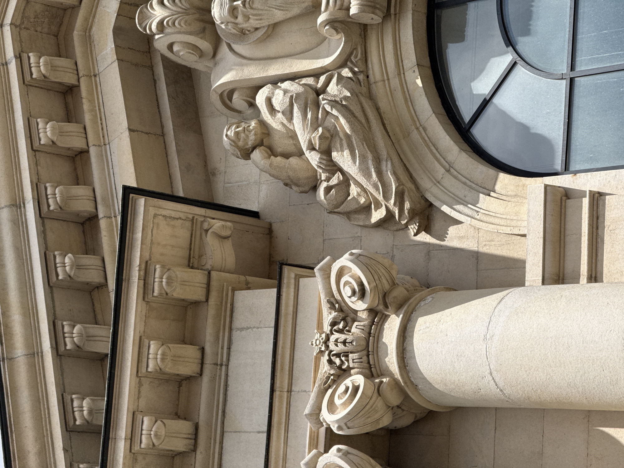 The image shows architectural details of a building facade. The building is constructed of light-colored stone and features a column with a decorative capital, a window with a circular design, and sculpted figures. The sculpted figures appear to depict people in flowing robes. The building's facade is heavily ornamented with classical motifs.
While it is difficult to pinpoint the exact location in Sofia, Bulgaria, without further information, the architectural style suggests it could be a government building or a cultural institution with classical influences.