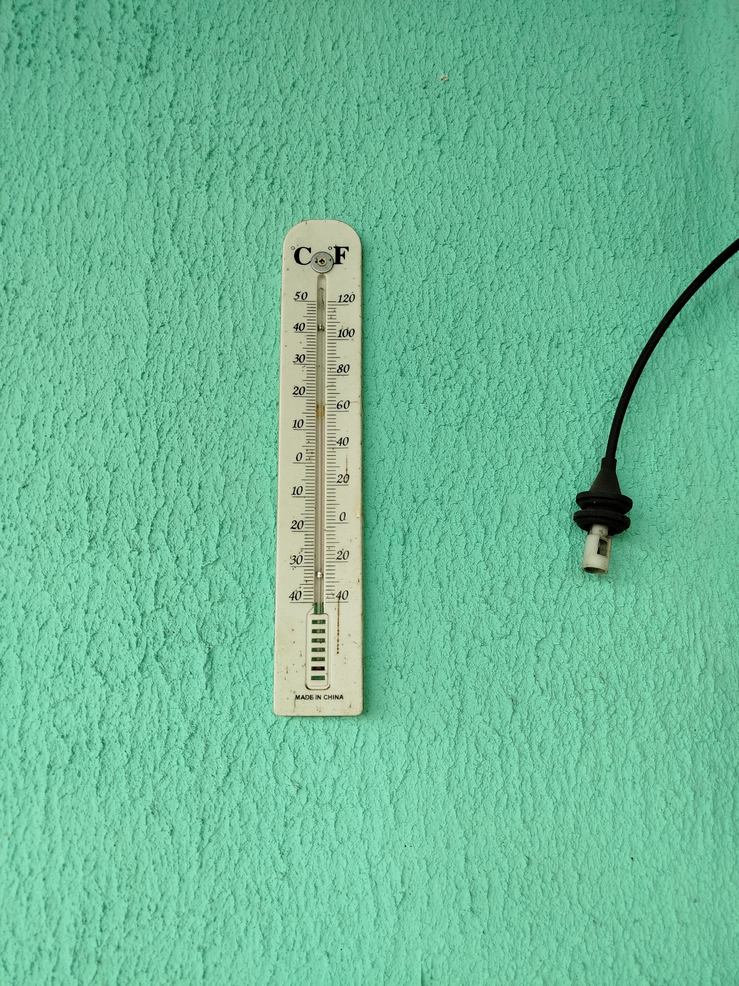An outdoor thermometer is mounted on a textured, light green wall. The thermometer is white and displays both Celsius and Fahrenheit scales. The mercury level in the thermometer indicates a temperature between 30 and 40 degrees Fahrenheit, and between 0 and 10 degrees Celsius. To the right of the thermometer, a black cable with a white plastic connector protrudes from the wall. The environment appears to be an outdoor setting, possibly a building exterior, in Крывасельскі сельскі Савет, Belarus.