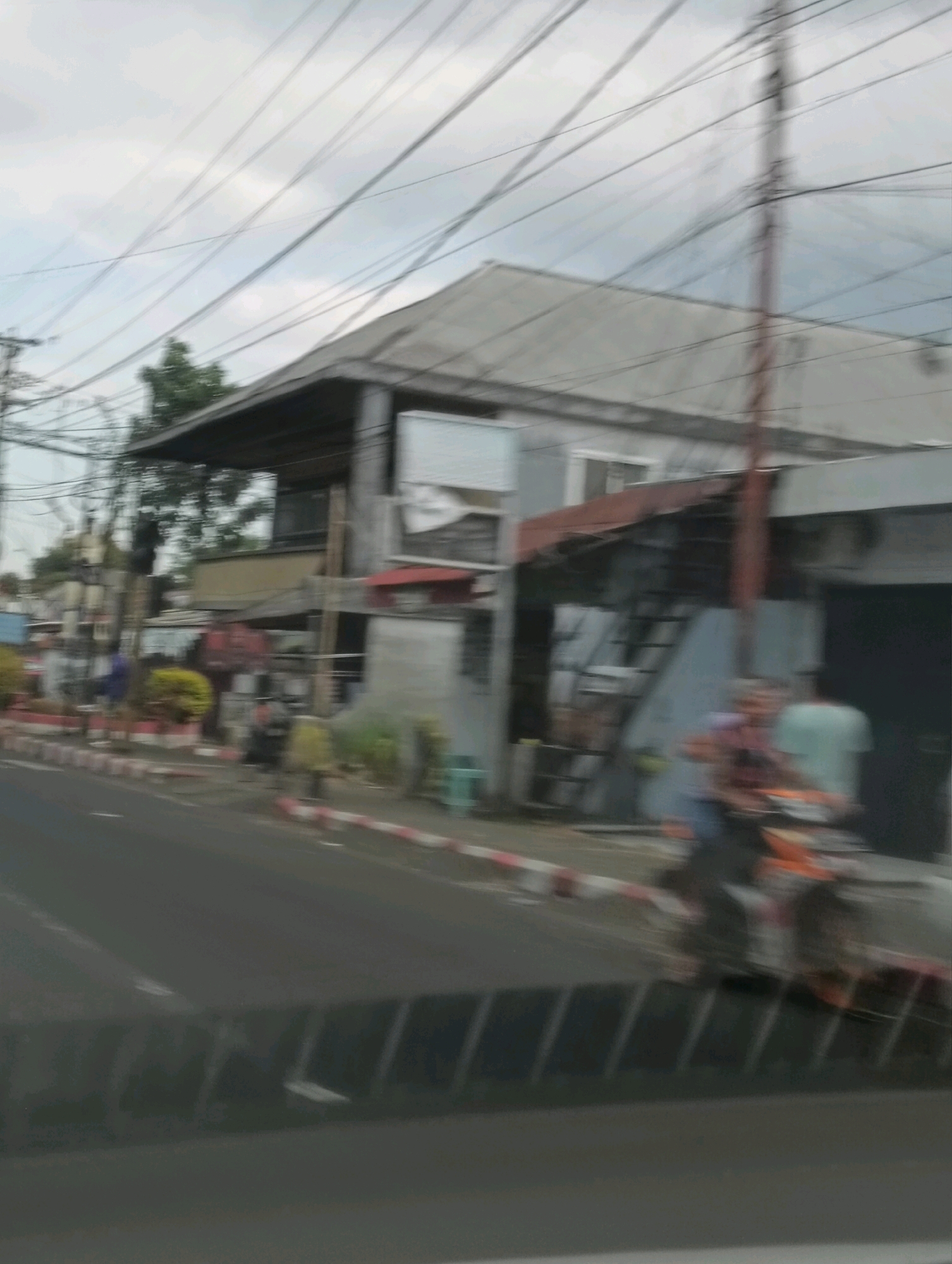 A street scene in Tomohon, Indonesia, under an overcast sky, features a multi-story building with a weathered facade and a corrugated metal roof. A complex network of overhead electrical wires dominates the upper portion of the image, crisscrossing the sky. In front of the building, a sidewalk with red and white striped barriers separates the pedestrian area from the asphalt road. Several people are visible on the sidewalk, with some appearing to be standing near small businesses or stalls. One individual is seen riding a motorcycle, which is mostly obscured by motion blur. The overall impression is one of an urban environment with signs of daily life.