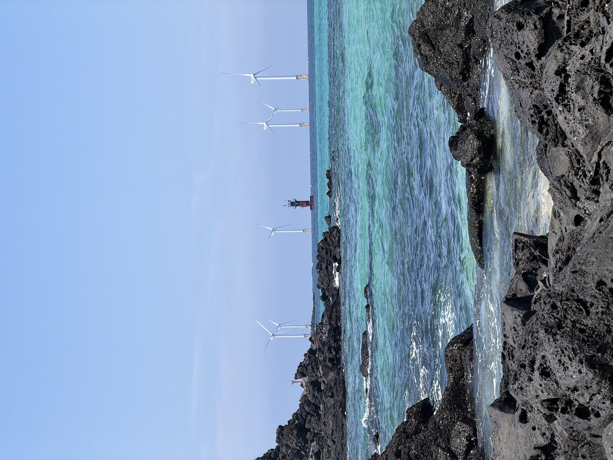 A wide, clear, bright blue sky stretches above a tranquil, turquoise ocean. In the middle ground, several tall, white wind turbines stand in the water, their blades still. A small, red and black lighthouse is also visible in the water. The foreground of the image features a rocky coastline made of dark, porous volcanic rock. Some of the rocks are partially submerged in the clear water, with small waves lapping at their edges. The scene appears to be a scenic coastal area, likely an offshore wind farm visible from the shore. This location is consistent with coastal areas around Jeju Island, South Korea, known for its volcanic landscapes and wind energy projects. There are no people or animals visible in the image.