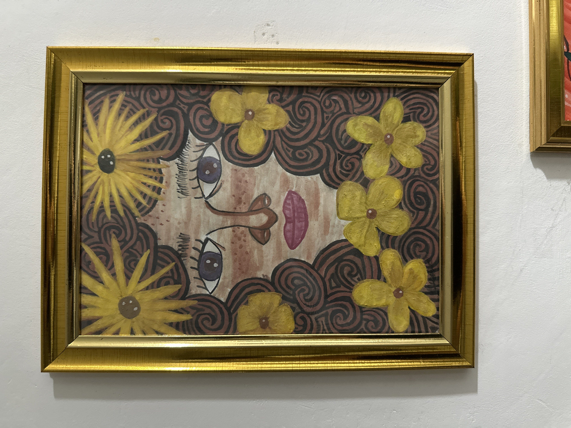 A framed artwork hangs on a white wall. The frame is gold-colored and ornate. The artwork depicts a stylized face with large eyes, a pink mouth, and brown features against a background of brown spirals. Yellow flowers are scattered around the face. The location is in Davao City, Philippines.