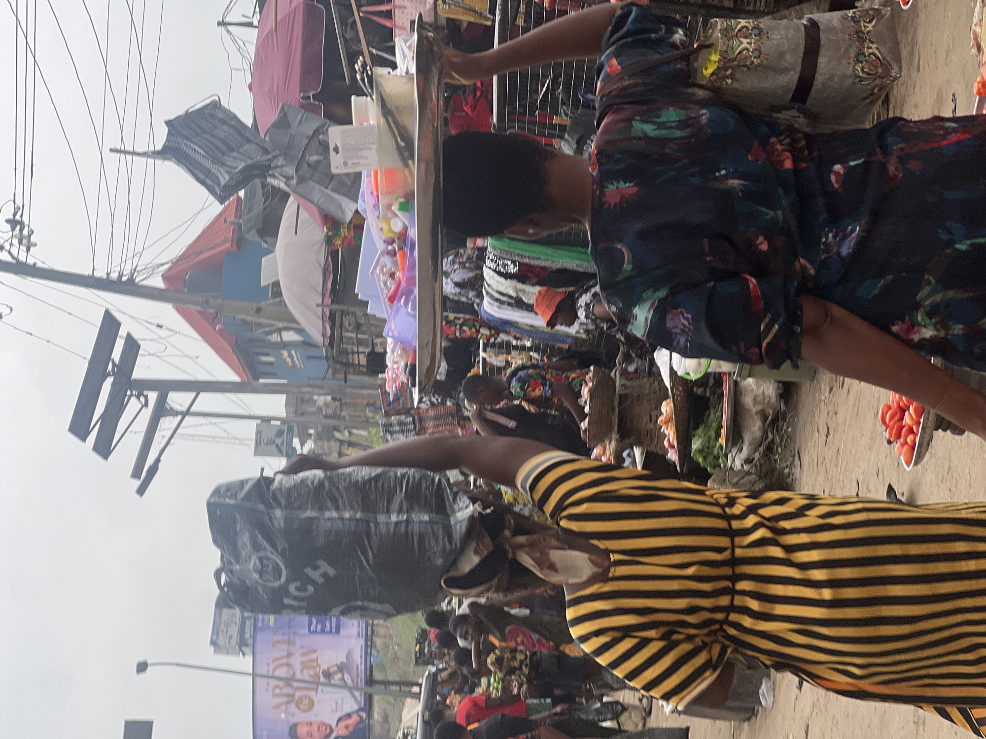 The image depicts a bustling market scene in Nkpor, Nigeria, under an overcast sky. In the foreground, two women are central figures. The woman on the left, wearing a vibrant yellow and black striped dress, carries a large black plastic bag balanced on her head, partially obscuring her face. To her right, another woman, her back to the camera, wears a dark, multi-colored patterned top and carries a silver tray laden with various items, including plastic containers and what appear to be cosmetics, balanced on her head. Her left arm is raised, steadying the tray.
The background reveals a crowded marketplace with numerous stalls and people. Stalls are covered with tarpaulins and umbrellas in shades of red and maroon, providing shade for vendors and shoppers. Various goods are visible, including produce like tomatoes displayed in baskets and on trays, and fabrics or clothing hanging from stalls. Electrical wires crisscross overhead, typical of urban Nigerian settings. A large billboard advertising "ABOVE THE LAW" with a man in a suit is visible in the distance. The ground is unpaved, suggesting a dirt or sand surface common in such open-air markets. The overall impression is one of lively commerce and everyday life in a Nigerian city.