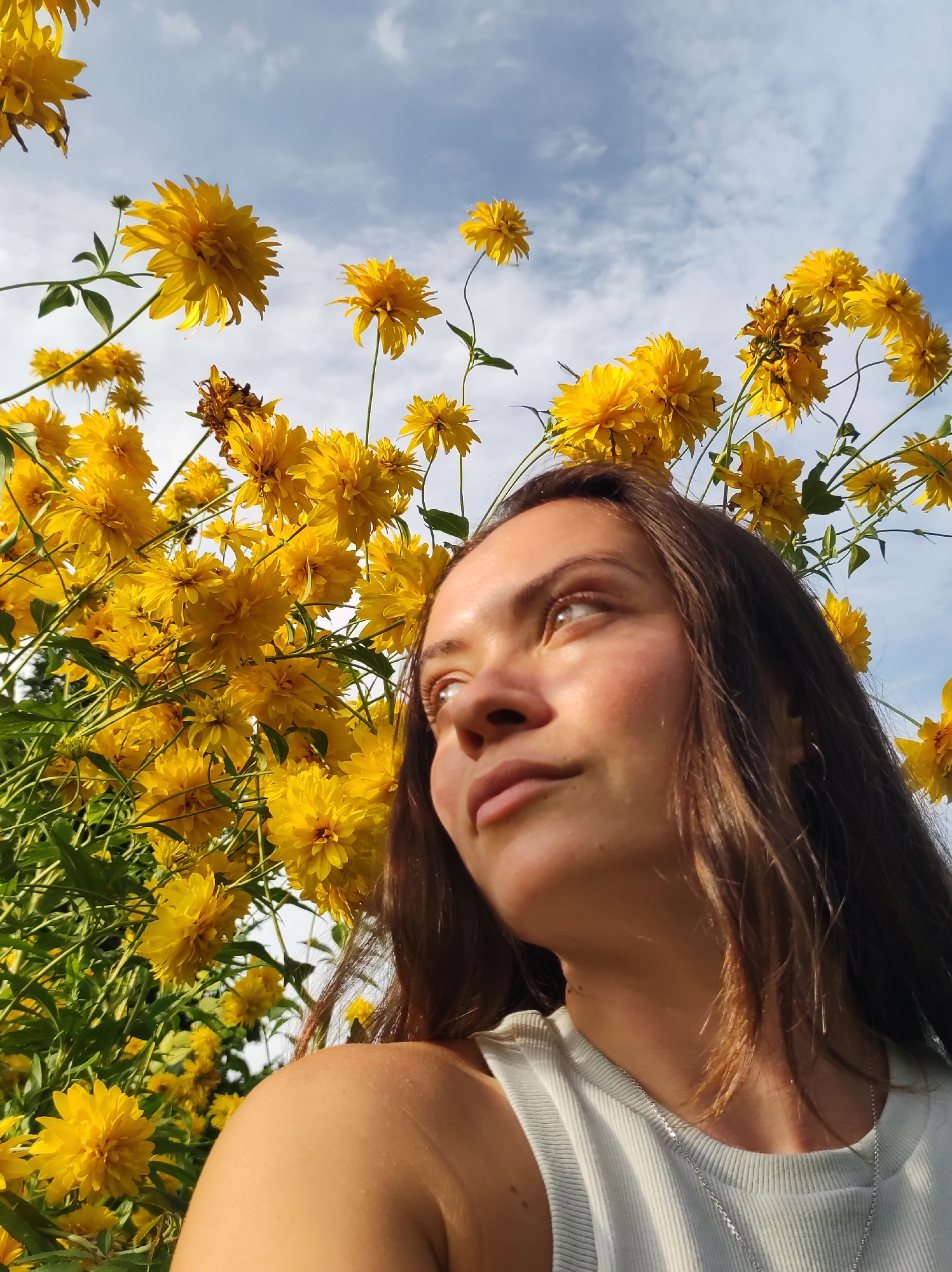 A young woman is captured in a low-angle, medium shot, looking upwards towards a bright, partly cloudy sky, with her right side of her face filling the frame. She has long, brown hair, and is wearing a light-colored, ribbed tank top with a silver necklace. She is positioned in front of a dense cluster of bright yellow, pom-pom-like flowers, which are also reaching towards the sky. The background consists of blue sky with scattered white clouds. The scene appears to be outdoors, in a natural setting, possibly a garden or park in the city of Zavod Mikhailovskii, Russia, given the flowers and the bright daylight.