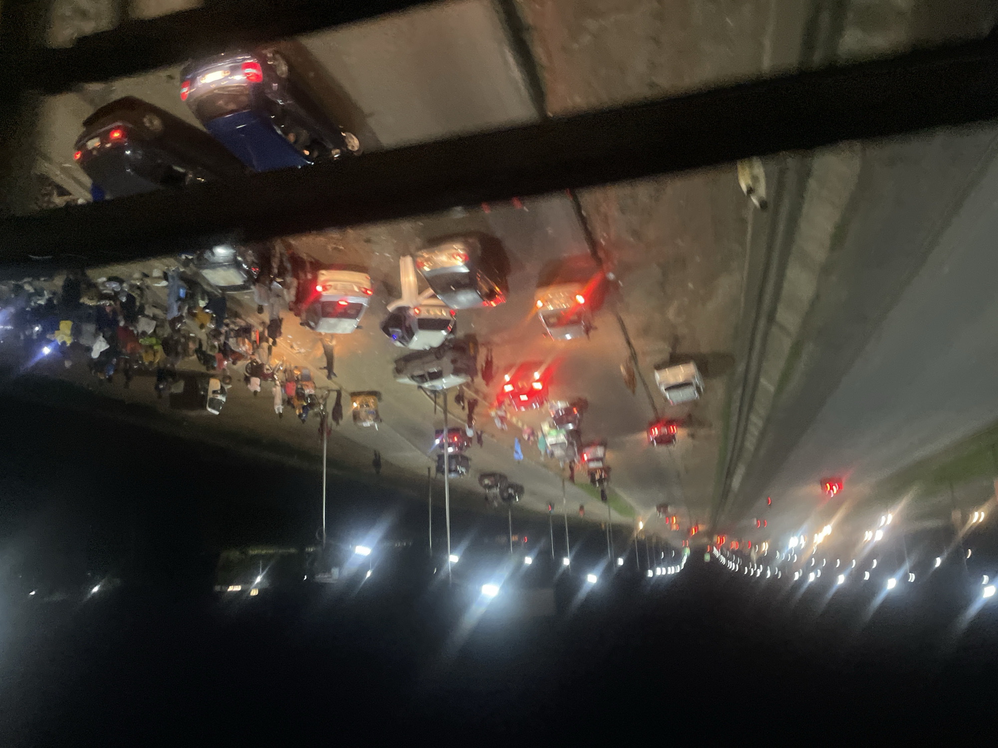 This media file captures a nighttime scene, likely in Sauka, Nigeria, that appears to be a car accident or a major disruption on a road. The image is taken from an elevated perspective, looking down on a highway or a large road.
The primary subjects are numerous vehicles, many with their brake lights illuminated, suggesting they have stopped abruptly. Several cars are visible, some appearing to be involved in a collision or stopped due to an obstruction. In the foreground and middle ground, there are groups of people gathered on the road, some appearing to be onlookers or emergency responders. They are illuminated by the lights of the vehicles and streetlights.
The scene is set at night, as indicated by the darkness and the prominent vehicle lights. The presence of streetlights and the general road infrastructure suggest an urban or semi-urban environment. The activity taking place seems to be a chaotic halt of traffic, with people congregating on the road. It's difficult to ascertain specific weather conditions, but the image appears clear. There are no discernible emotions that can be clearly identified due to the distance and low light.
Notable details include the presence of what appear to be utility poles with lights, illuminating sections of the road. The sheer number of people and vehicles suggests a significant event. The image is somewhat blurry, possibly due to low light conditions, movement, or the quality of the recording device. There is no visible text in the image.