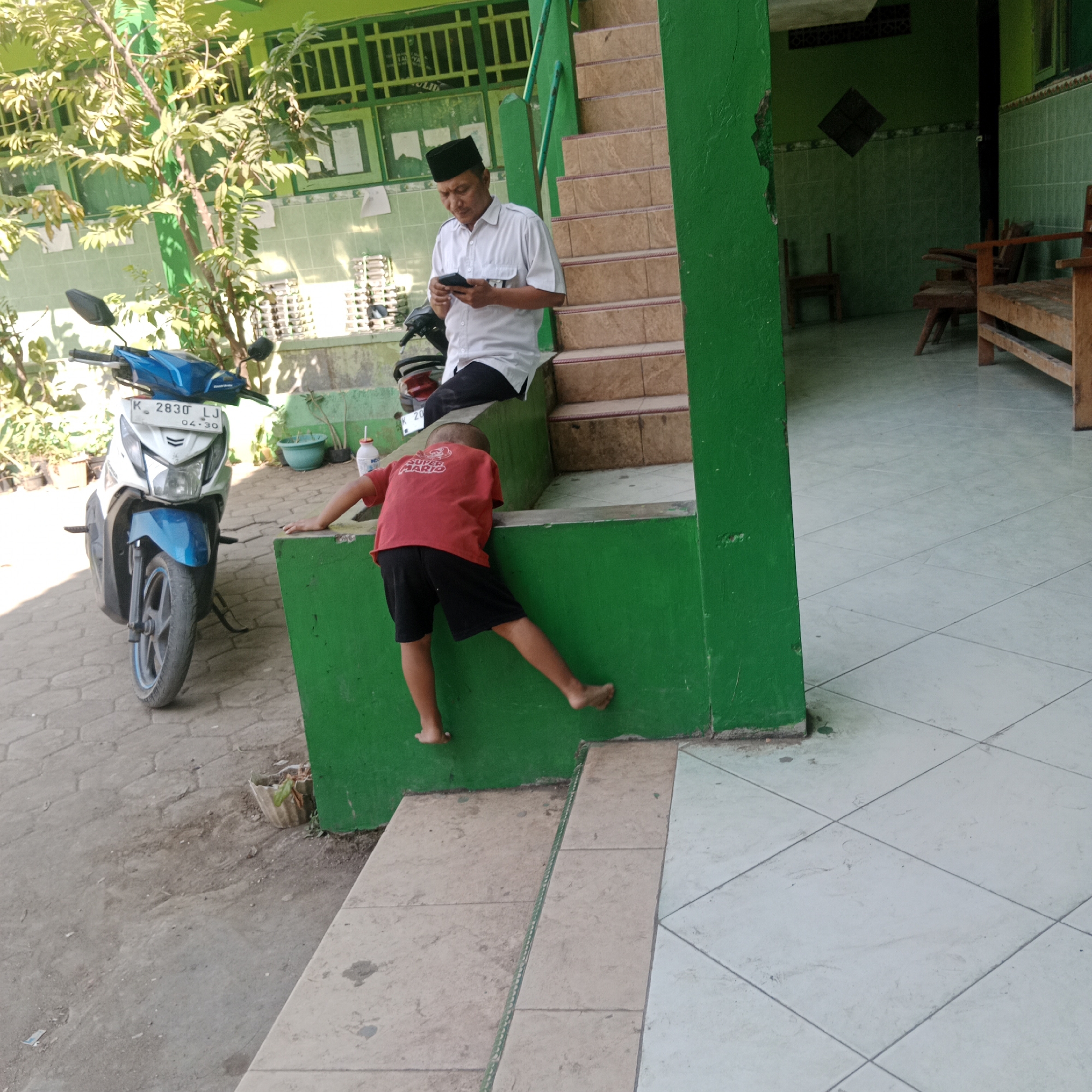 In the city of Tegowanu Kulon, Indonesia, a man wearing a white shirt and a black peci (cap) is seated on a green ledge, looking at his mobile phone. Next to him, a young boy in a red t-shirt with "Super Mario" printed on it and black shorts is climbing or playing on the green ledge. A blue and white motorbike is parked to the left of the man. The background shows a building with green tiled walls and a set of brown tiled stairs. Trees with green leaves are visible on the left side of the image. The ground is paved with concrete blocks.