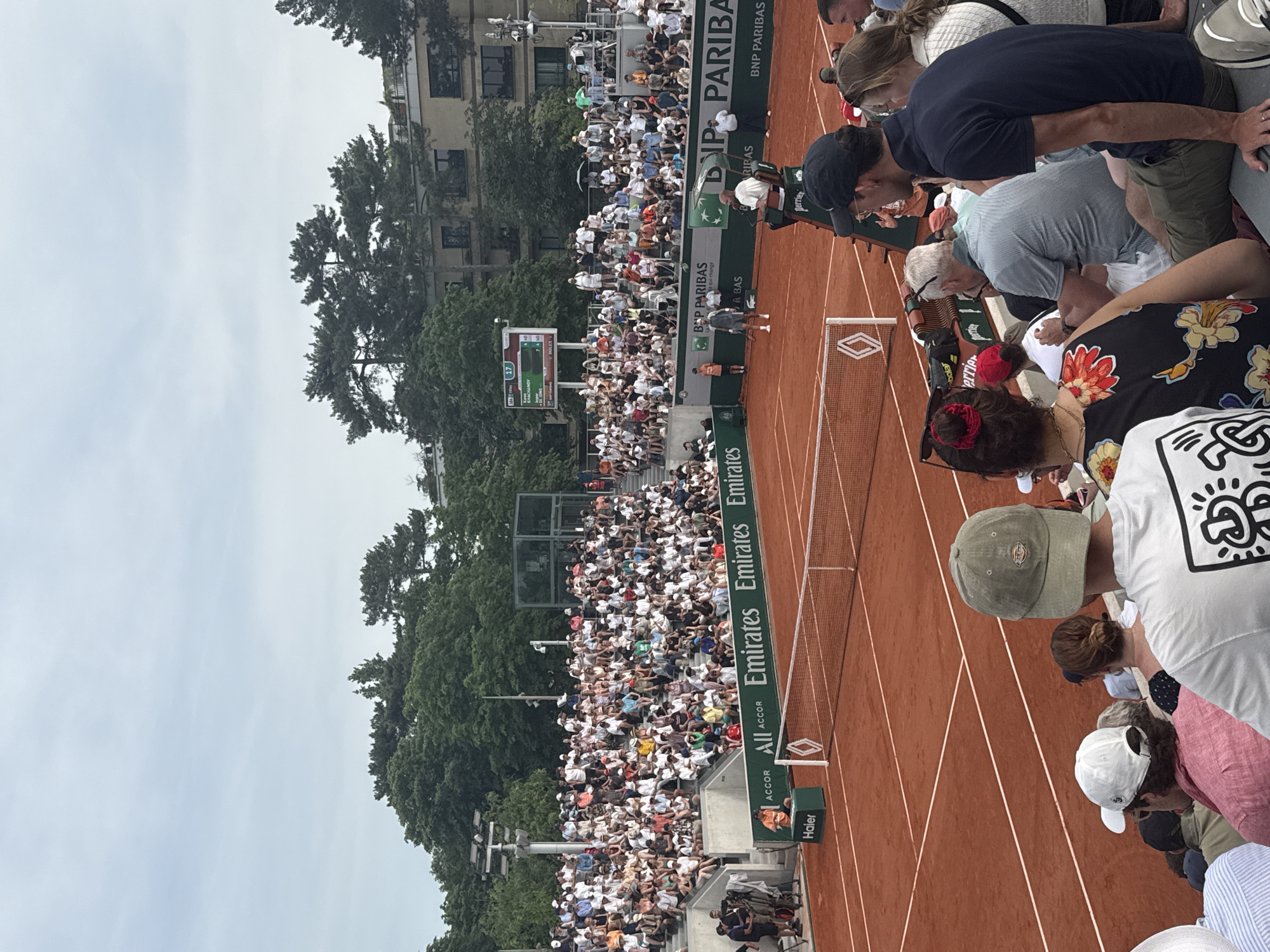 This outdoor image depicts a tennis match taking place at Roland Garros in Paris, France. A clay court is visible with white lines marking the boundaries, and a net is stretched across the center. The stands are filled with spectators, many of whom are looking towards the court. Advertising banners for companies such as Emirates, Accor, and BNP Paribas are visible along the edges of the court. In the background, trees and buildings can be seen. The weather appears to be overcast.