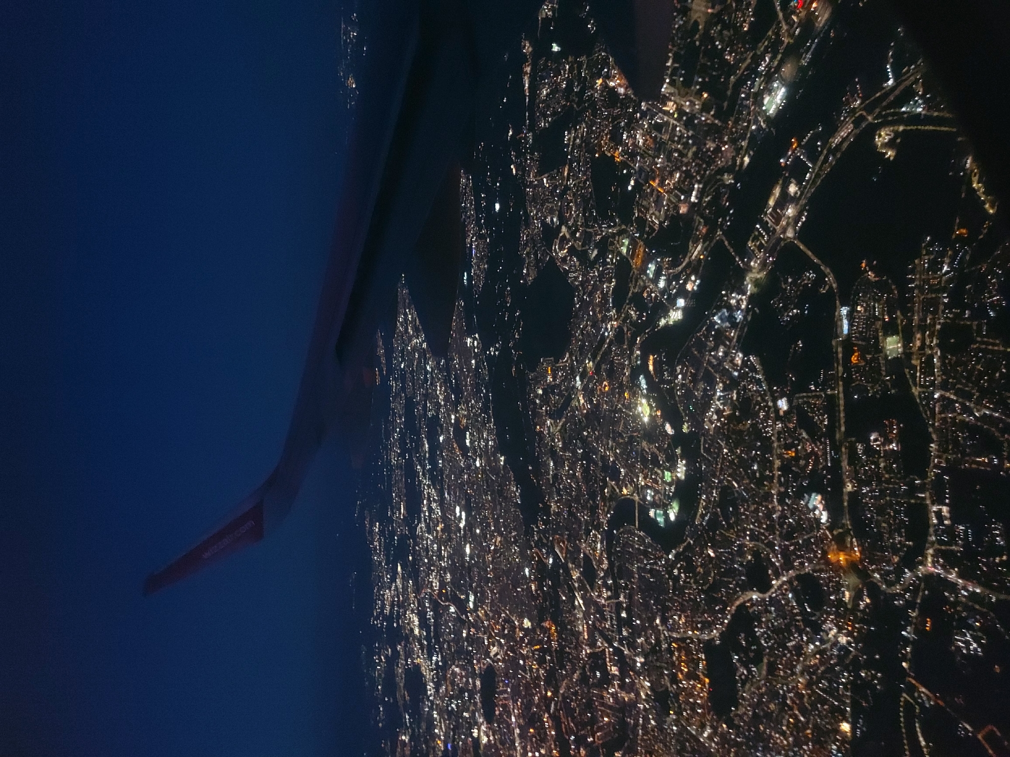 The image depicts a nighttime view of the city of Fiumicino, Italy, from the perspective of an airplane window. Visible is the wing of the plane and, below, a sprawling urban landscape illuminated by streetlights and building lights. The city's grid-like pattern of streets is discernible, with brighter clusters of lights indicating possibly commercial or densely populated areas. The wing of the plane is branded with "wizzair.com." The environment outside the city is dark blue.
