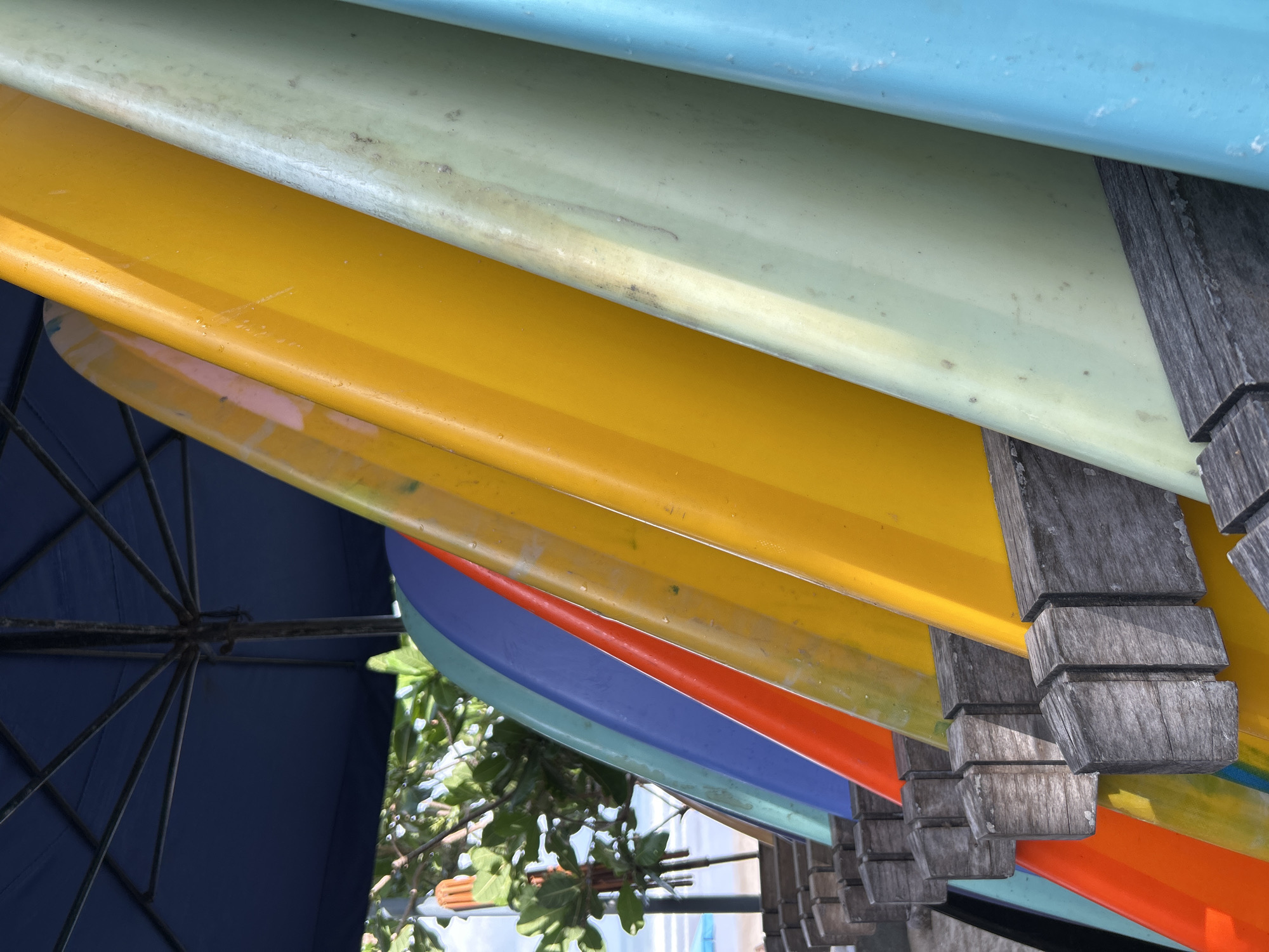An eye-level, close-up shot captures a rack of colorful surfboards lined up under a dark blue umbrella, likely at a surf school or rental shop near the beach in Kuta, Bali, Indonesia. The surfboards, varying in vibrant hues such as orange, yellow, light green, and bright blue, are angled upwards and secured in weathered wooden cradles. Sunlight highlights the smooth, glossy surfaces of the boards, revealing subtle scuffs and water marks. In the background, out of focus, hints of lush green foliage and a sliver of the ocean with distant umbrellas can be discerned, suggesting a typical beach setting. The scene is static, with no people or ongoing activity visible, focusing entirely on the organized display of surf equipment.