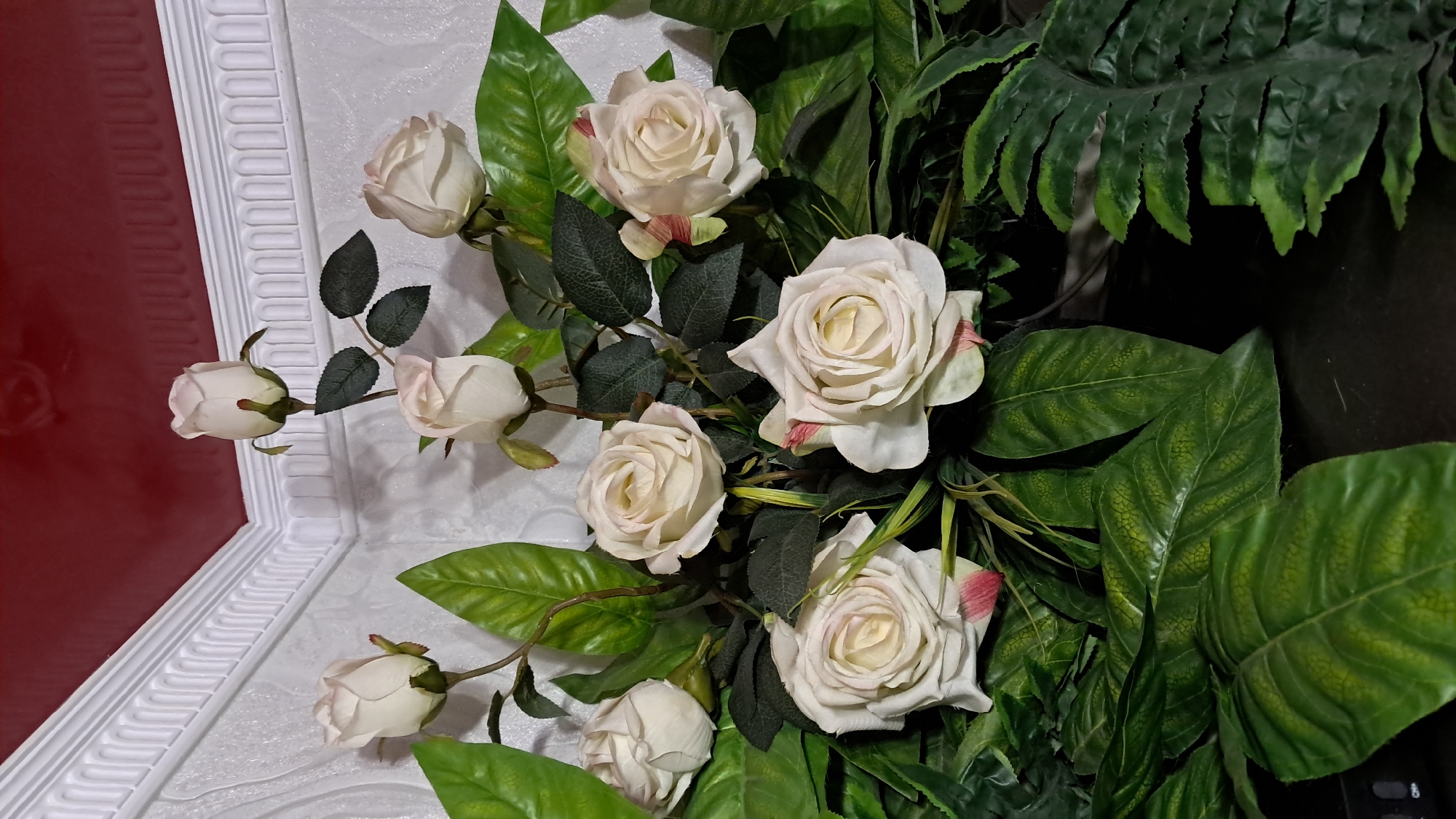 The image shows an arrangement of artificial cream-colored roses and green foliage. The roses are at various stages of bloom, with some fully open and others still budded. The leaves are a deep green, with some appearing glossy and others matte. The arrangement is placed against a textured white wall, with white decorative molding visible at the top. Above the molding, a red ceiling is visible. The lighting is artificial and casts shadows on the wall. The scene is static, with no people or actions depicted. The location is unspecified, but the request mentions Vinnytsia, Ukraine, which is a city. The arrangement appears to be decorative, possibly for an interior setting.