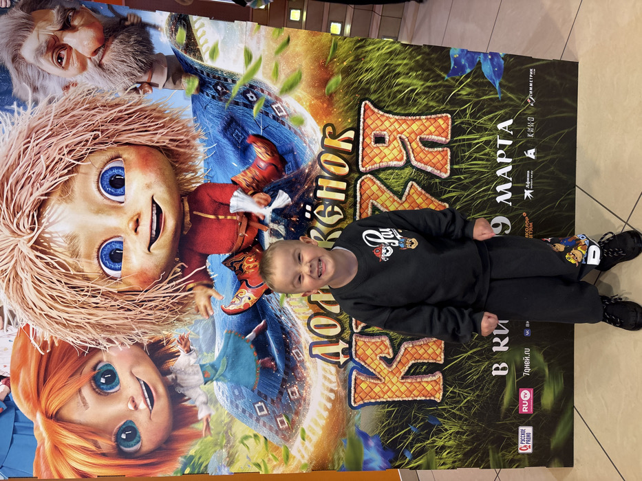 A young male child, estimated to be between four and six years old, stands centered in the foreground, posing in front of a large movie poster. The child has light skin and short, light brown hair, and is captured mid-smile with his eyes slightly squinted and cheeks raised, indicating a cheerful expression. He is dressed in a black long-sleeved sweatshirt featuring a "Paw Patrol" logo on the left chest, along with matching black sweatpants adorned with multiple "Paw Patrol" characters and "PAW" text on the lower left leg. He wears black sneakers with white laces.
The background is dominated by a colorful, vertically oriented poster for the animated film "Домовёнок Кузя" (Kuzya the House Elf). The poster depicts three animated characters: a red-haired girl with blue eyes on the left, a blonde-haired boy with blue eyes in the center, and an elderly man with a white beard on the right. The central blonde-haired boy wears a red tunic and pointed boots and appears to be sitting on a blue, patterned flying carpet. The film title is prominently displayed in large, textured golden-brown Cyrillic letters: "ДОМОВЁНОК КУЗЯ." Below the title, text indicates "В КИНО С 7 МАРТА" (In cinemas from March 7th) and "9 МАРТА" (March 9th), along with various sponsor and media logos. The poster's lower portion depicts green grass and scattered leaves.
The scene takes place indoors, likely within a cinema lobby or entertainment complex in the city of Chaikovsky, Russia, given the size and nature of the movie advertisement. The floor is a light-colored tiled surface, and a dark wall or structural element is partially visible on the right side of the frame.