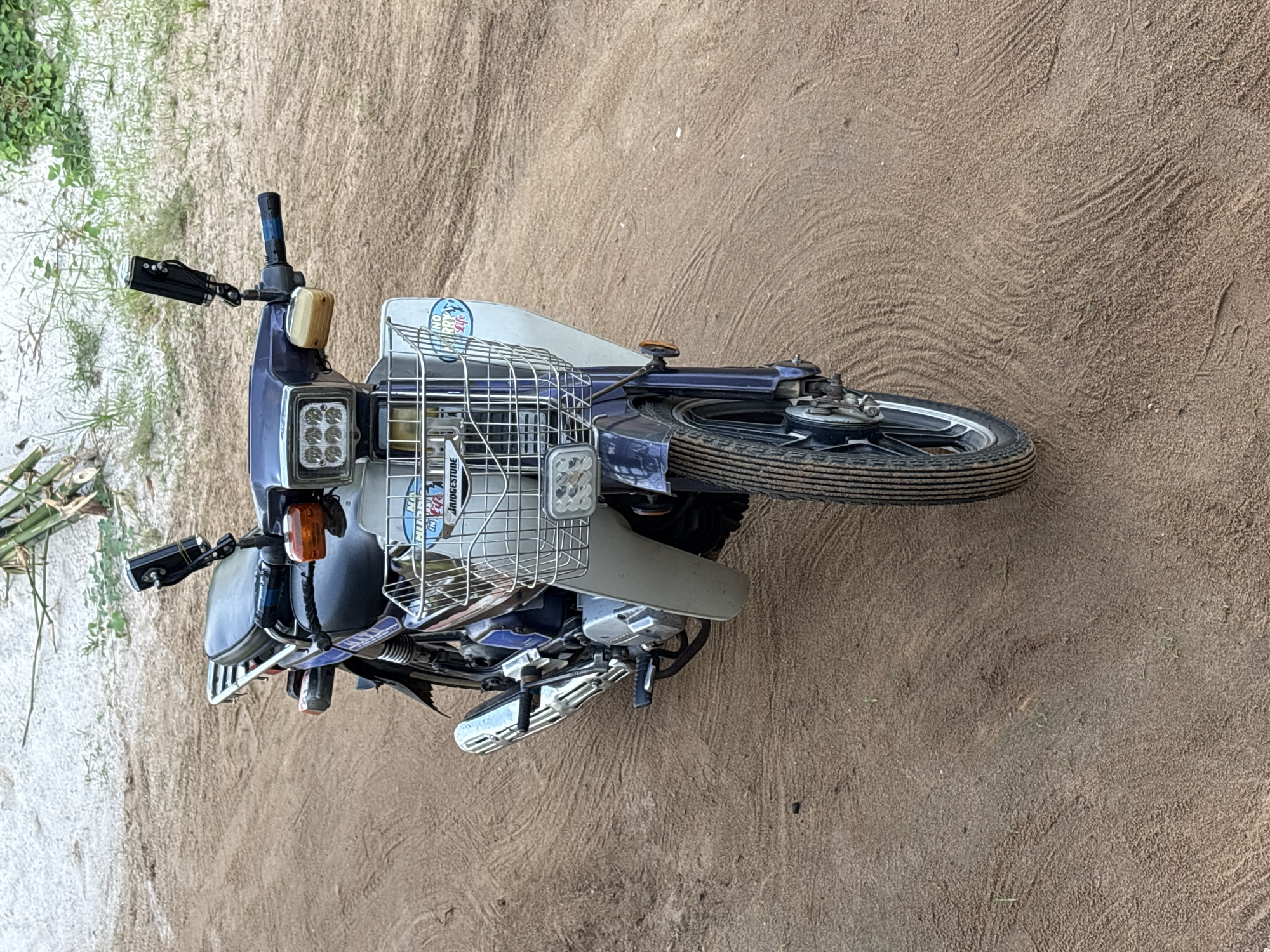A blue and silver motorcycle is parked on a patch of sand and grass in what appears to be Ikot Ekpene, Nigeria. The motorcycle has a metal basket attached to the front and additional headlights. The ground is sandy with patches of green vegetation.