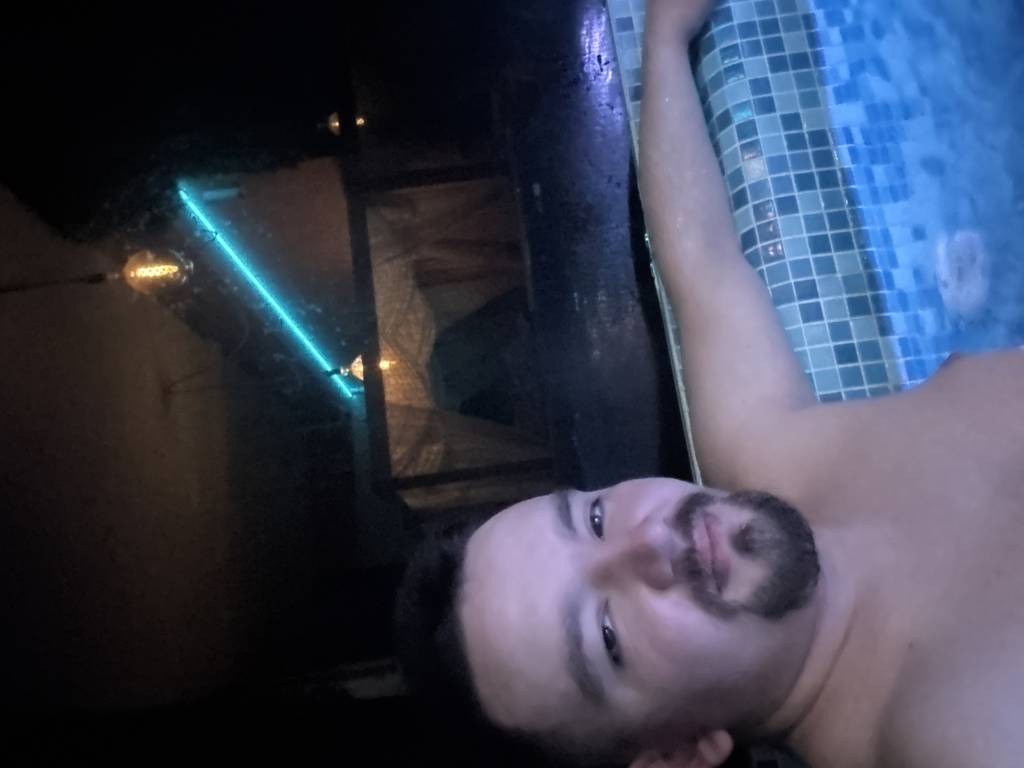 A man is lying near a blue-tiled pool or hot tub, possibly in Sofia, Bulgaria. He has dark hair and a dark goatee. He is facing the camera, with his head resting near the edge of the pool. The pool is lit with blue lights. There is a dim light in the background, and a neon light strip above it. The scene is dark and appears to be indoors. The exact location in Sofia, Bulgaria is unknown.