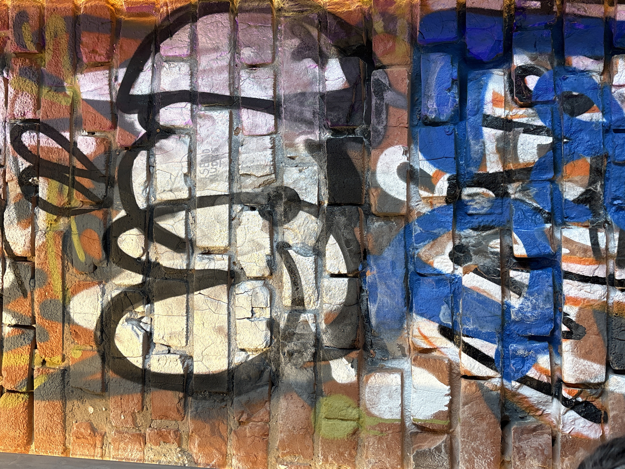 A brick wall covered with graffiti, located in Kazan, Russia. The graffiti consists of black and blue paint. The wall has a brick pattern with orange and white hues and a rugged, textured surface.